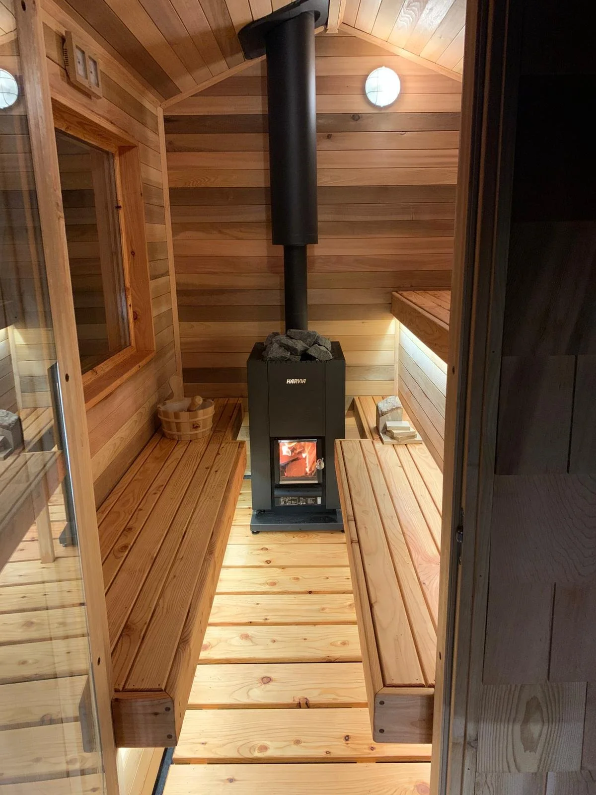 A small, cozy sauna with wooden benches on both sides and a wood-burning stove in the center, surrounded by a few sauna stones, with wooden walls and ceiling, and soft lighting.