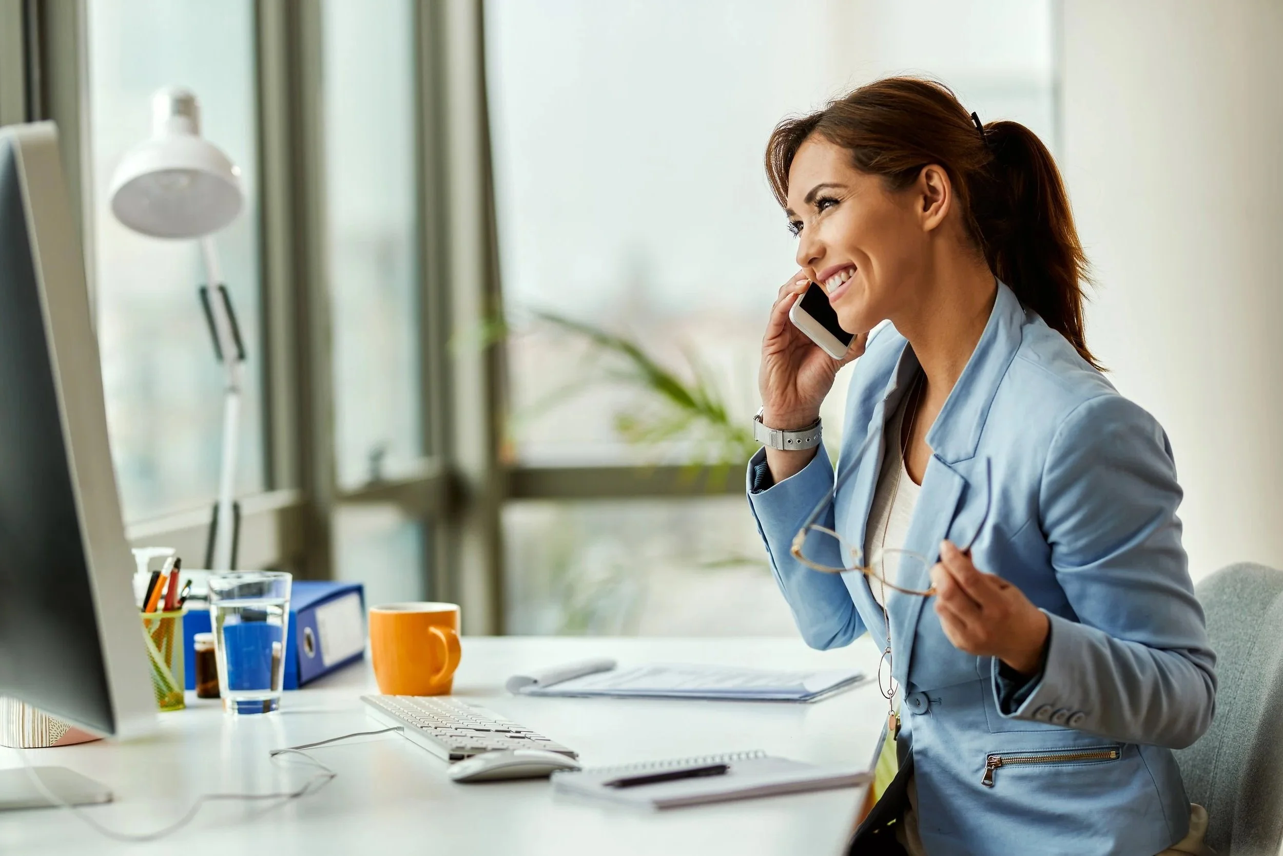 A woman sitting at a desk in an office, smiling while talking on a mobile phone and holding eyeglasses in her hand. The desk has a computer, glasses, a pen holder, a glass of water, an orange mug, and papers.