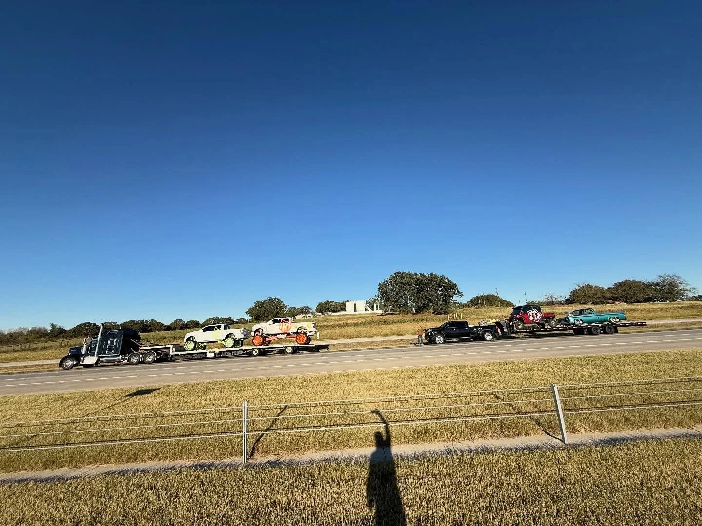 A black semi-truck hauling vintage cars on a flatbed trailer along a highway with grassy fields and trees under a clear blue sky.
