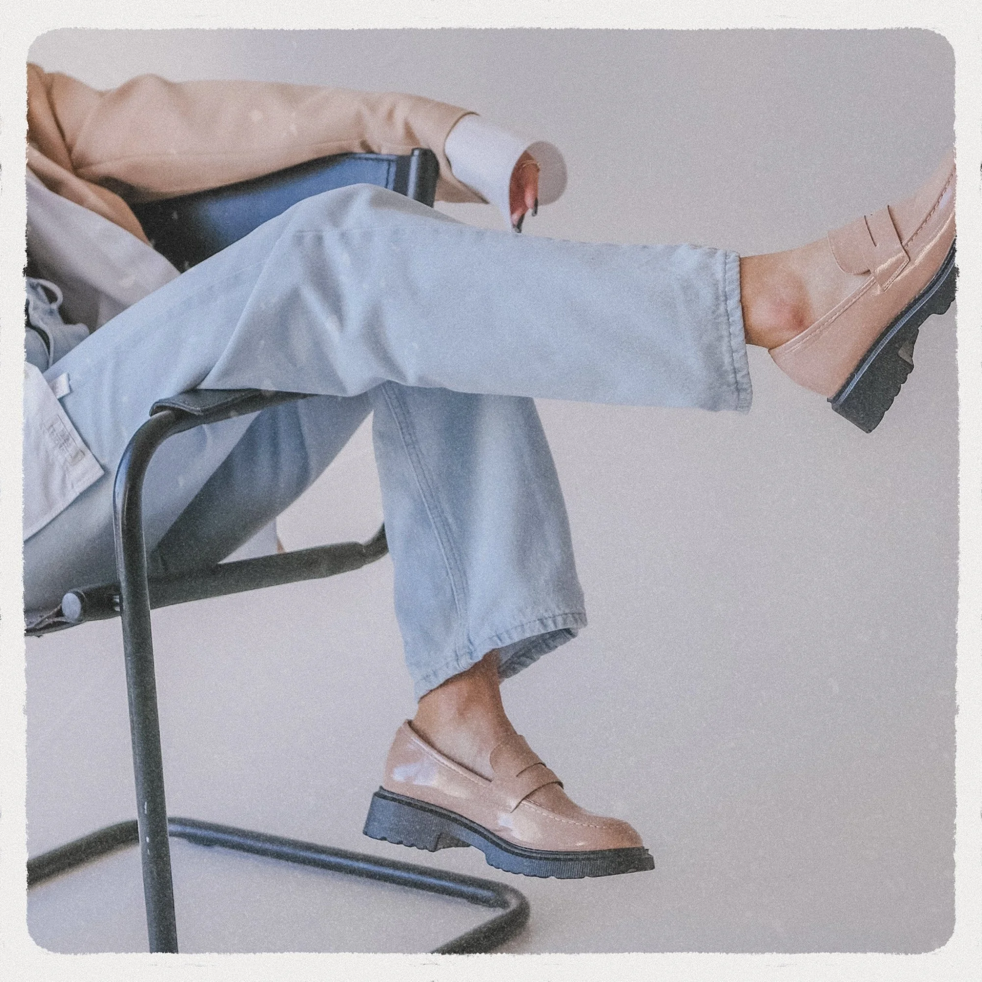 Person wearing tan shoes and light blue jeans sitting on a black chair against a neutral background.