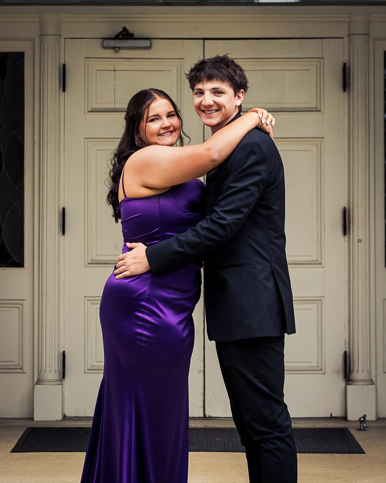 A young couple is dancing and smiling in front of a garage door. The woman is wearing a purple satin gown, and the man is dressed in a black suit. They are embracing each other affectionately.