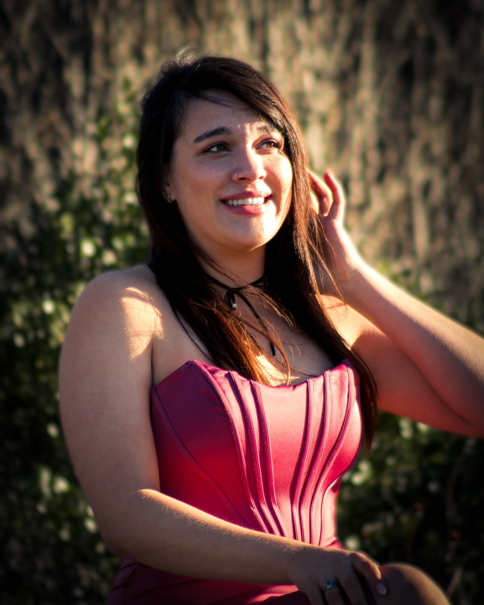 A woman with dark hair and freckles smiling outdoors in a pink dress with black accents and a choker necklace, touching her hair with one hand, with trees blurred in the background.