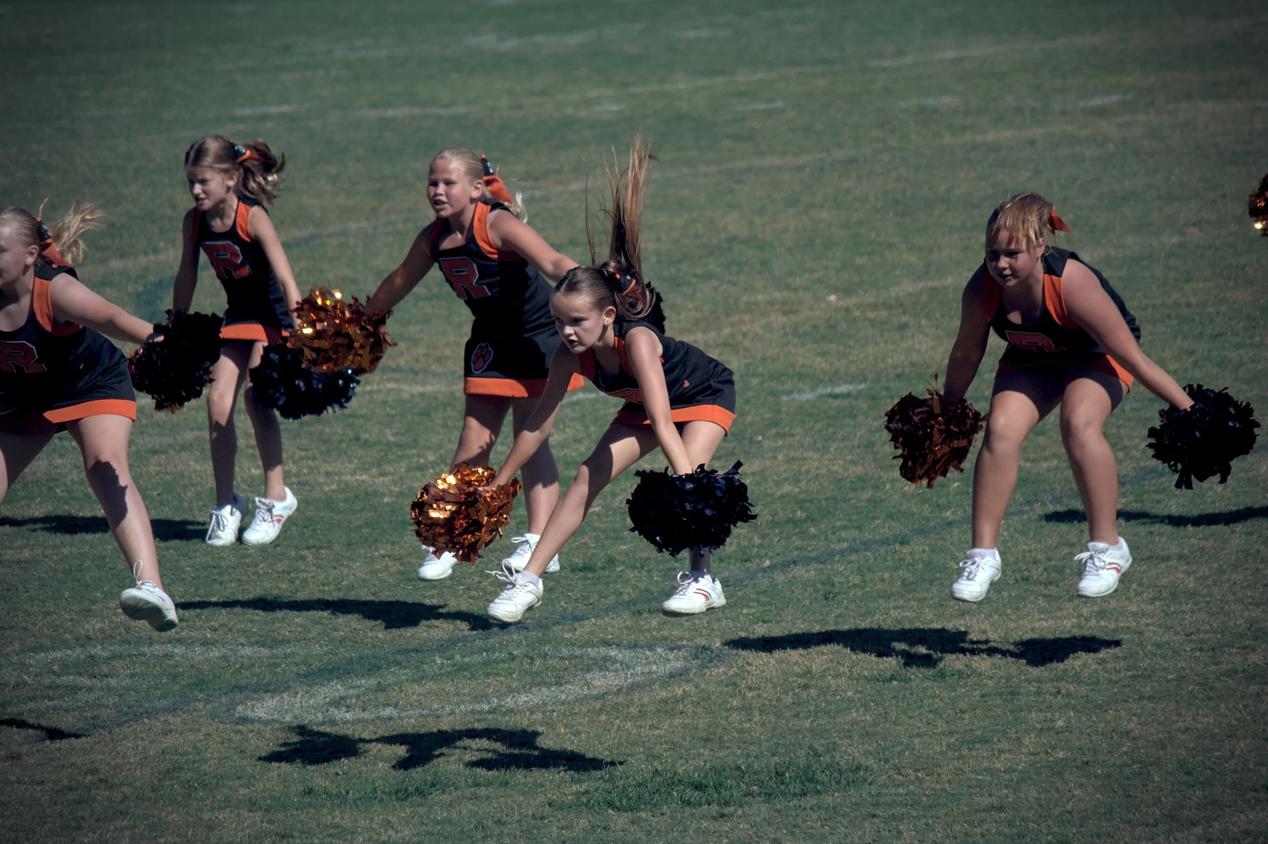 Five young cheerleaders in matching uniforms performing a cheer routine on a grassy field, holding pom-poms.