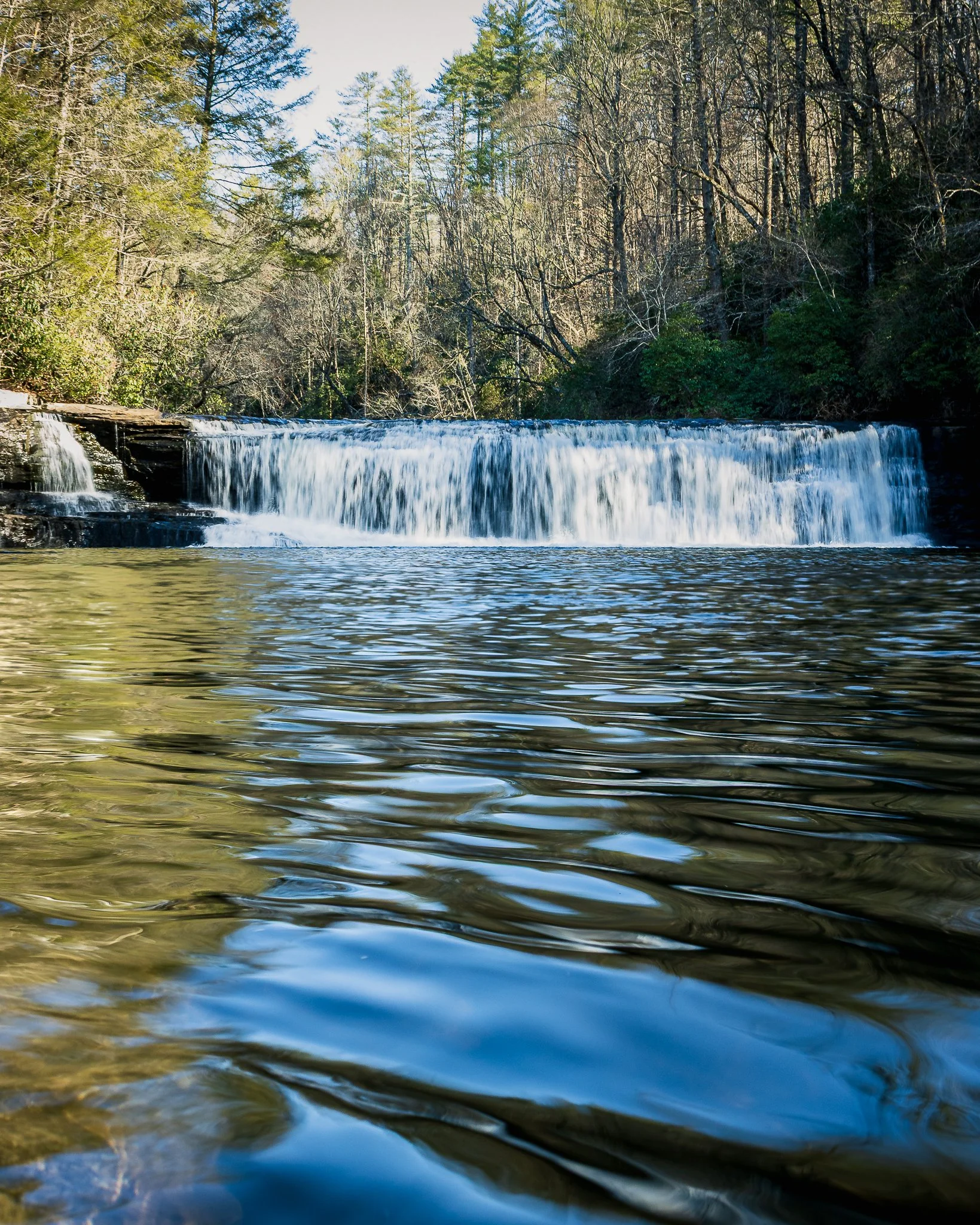 Calm river with a small waterfall at the edge, surrounded by a forest of tall trees, some with bare branches and some with green foliage, under a clear sky.