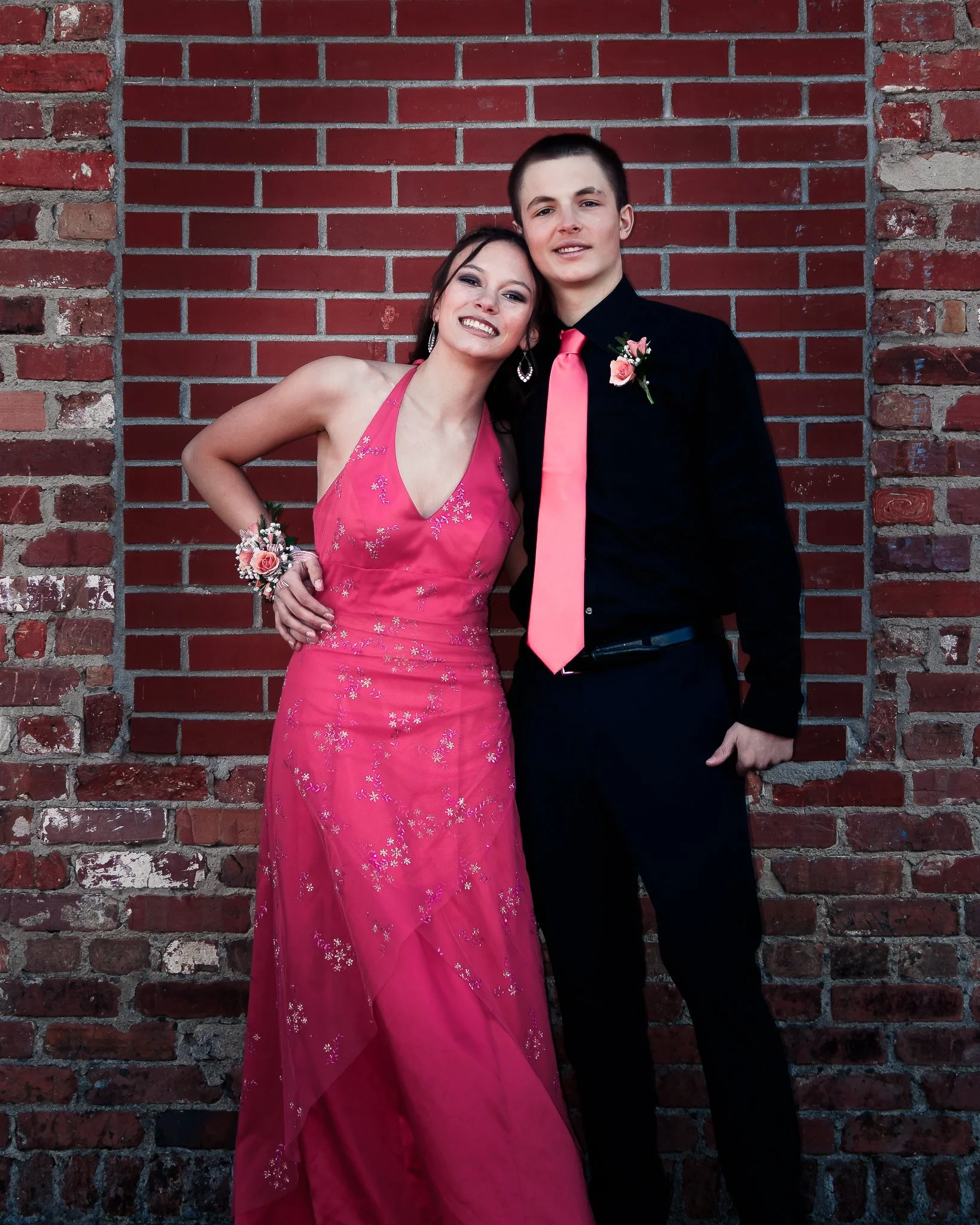 A young couple, dressed in formal attire, standing against a brick wall. The woman is wearing a pink gown with floral embroidery and jewelry, while the man is wearing a black shirt with a pink tie and boutonniere.
