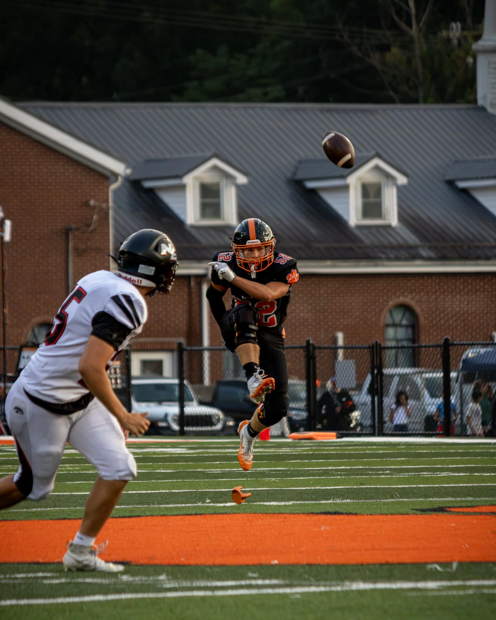 A football player in black and orange uniform jumping to catch a football during a game at a high school football field.