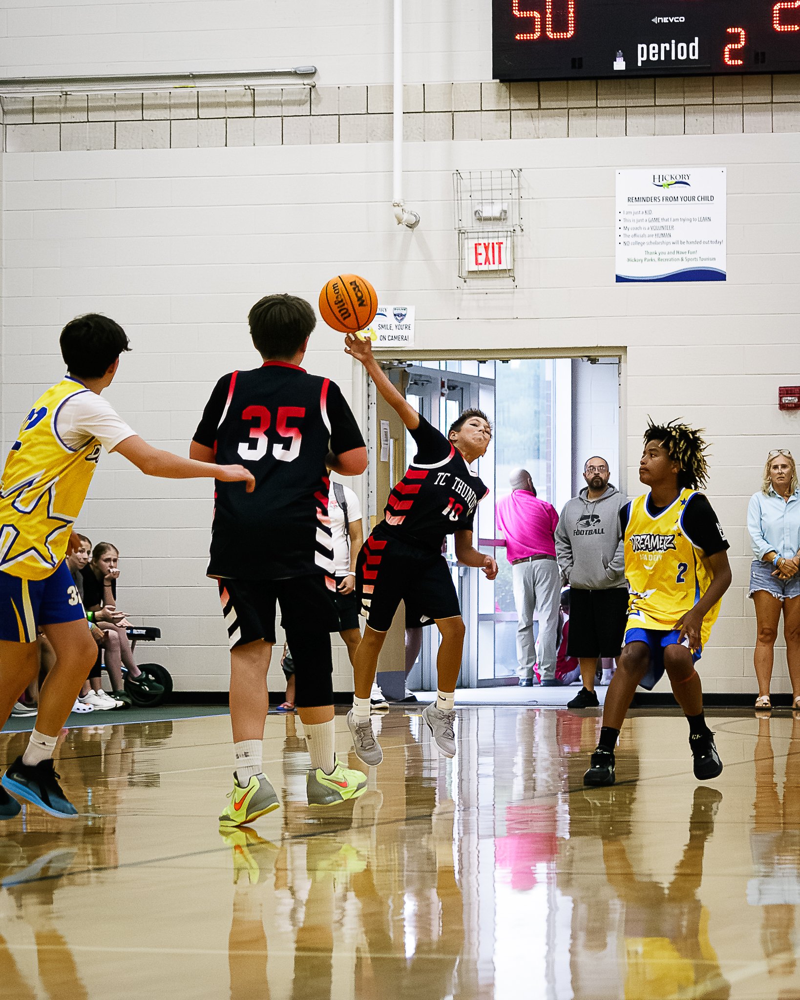 Kids playing basketball in a gymnasium, with some spectators watching.