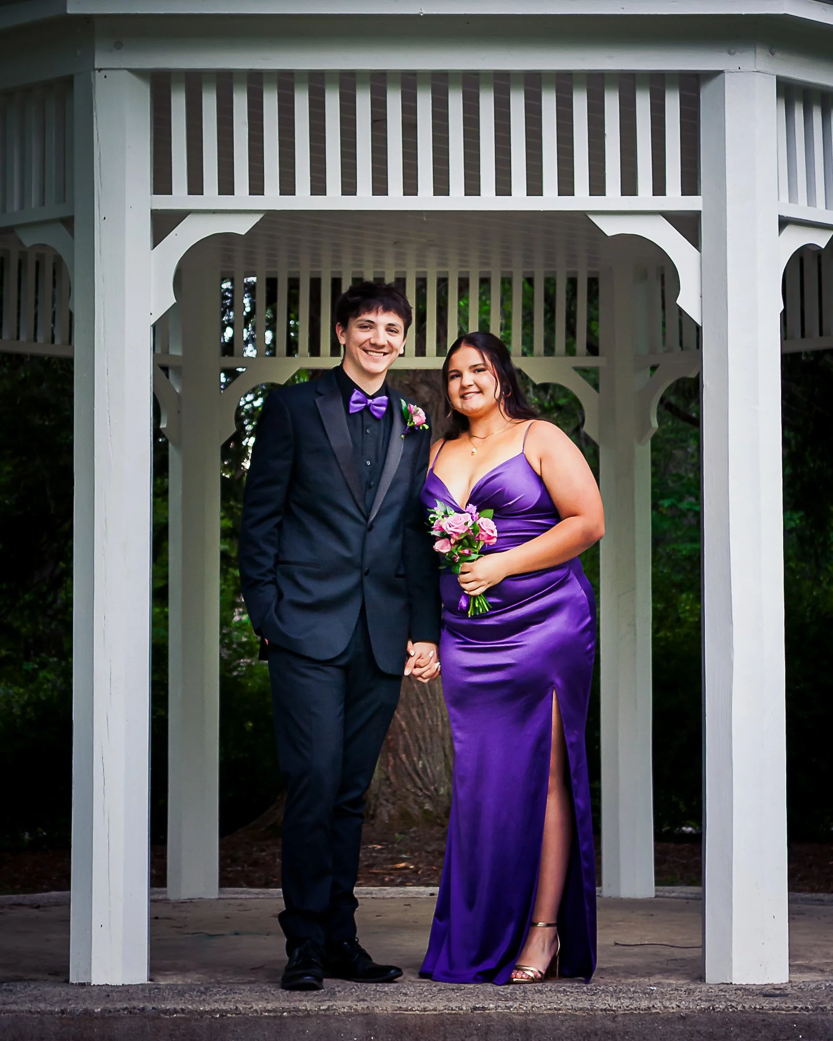 A young couple in formal attire holding hands and smiling, standing under a white wooden gazebo. The man is wearing a black suit with a purple bow tie and boutonniere, and the woman is wearing a purple satin gown and gold sandals, holding a bouquet o