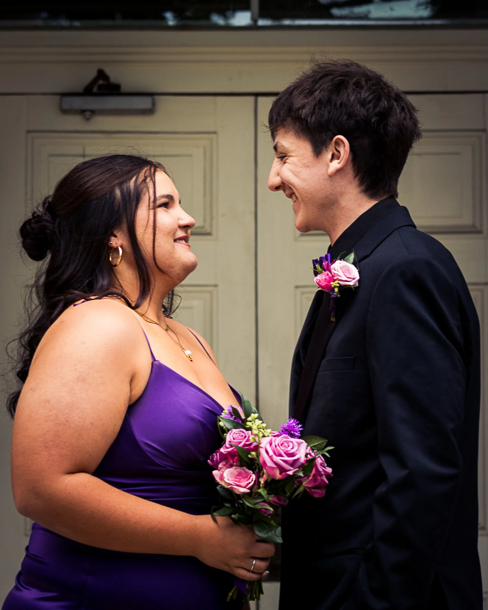 A woman and a man are smiling and looking into each other's eyes, with the woman holding a bouquet of pink and purple roses.