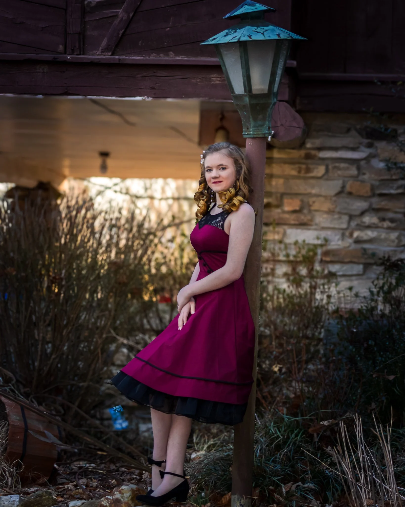 A young woman with curly hair, wearing a sleeveless magenta dress with black lace details and black shoes, leaning against a wooden lamp post outdoors.