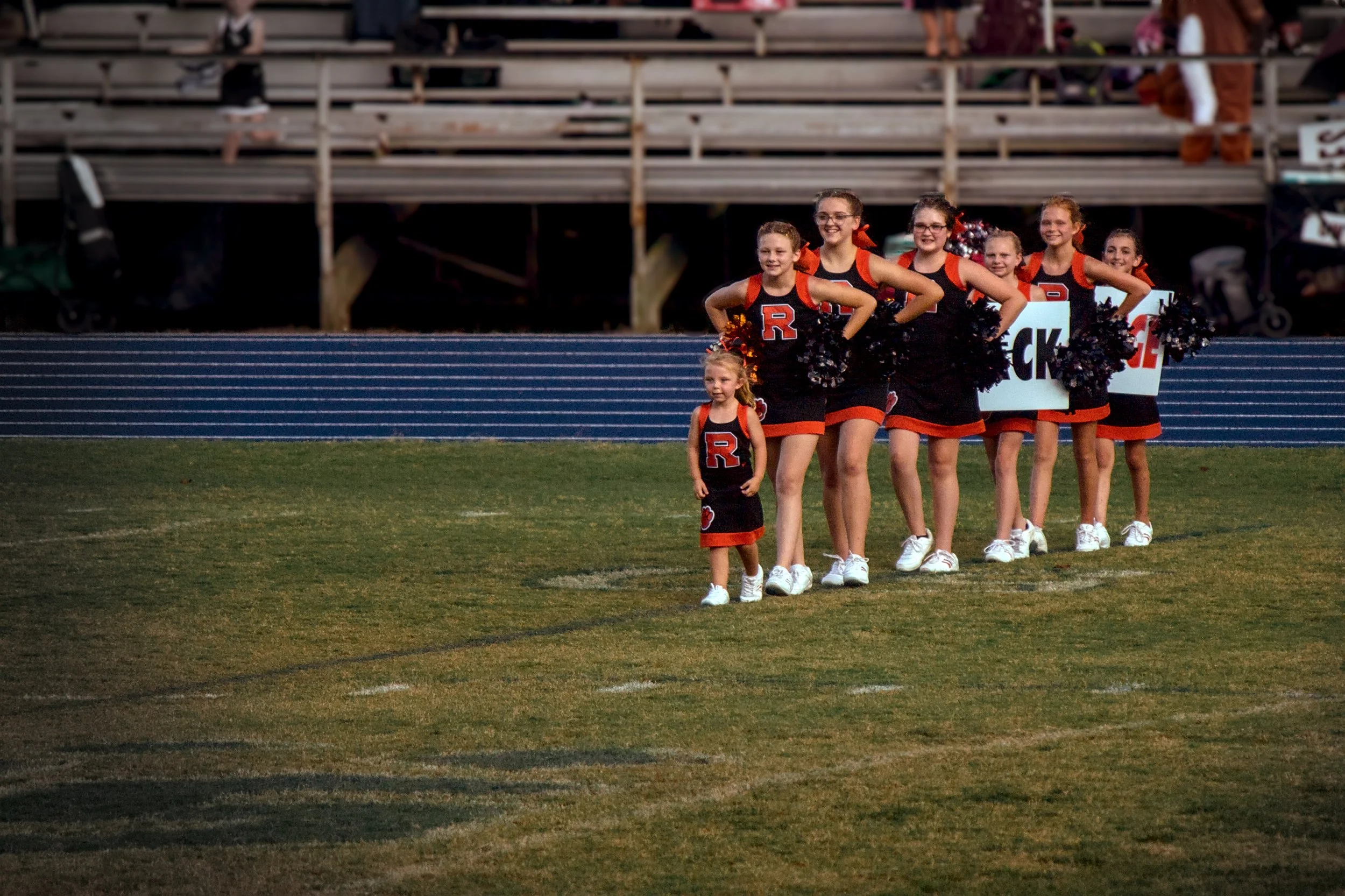 A group of young cheerleaders standing on a football field with a young girl in front, all wearing black and orange cheerleading uniforms with an 'R' on the front. They are holding black pom-poms and are smiling, with bleachers and spectators in the 