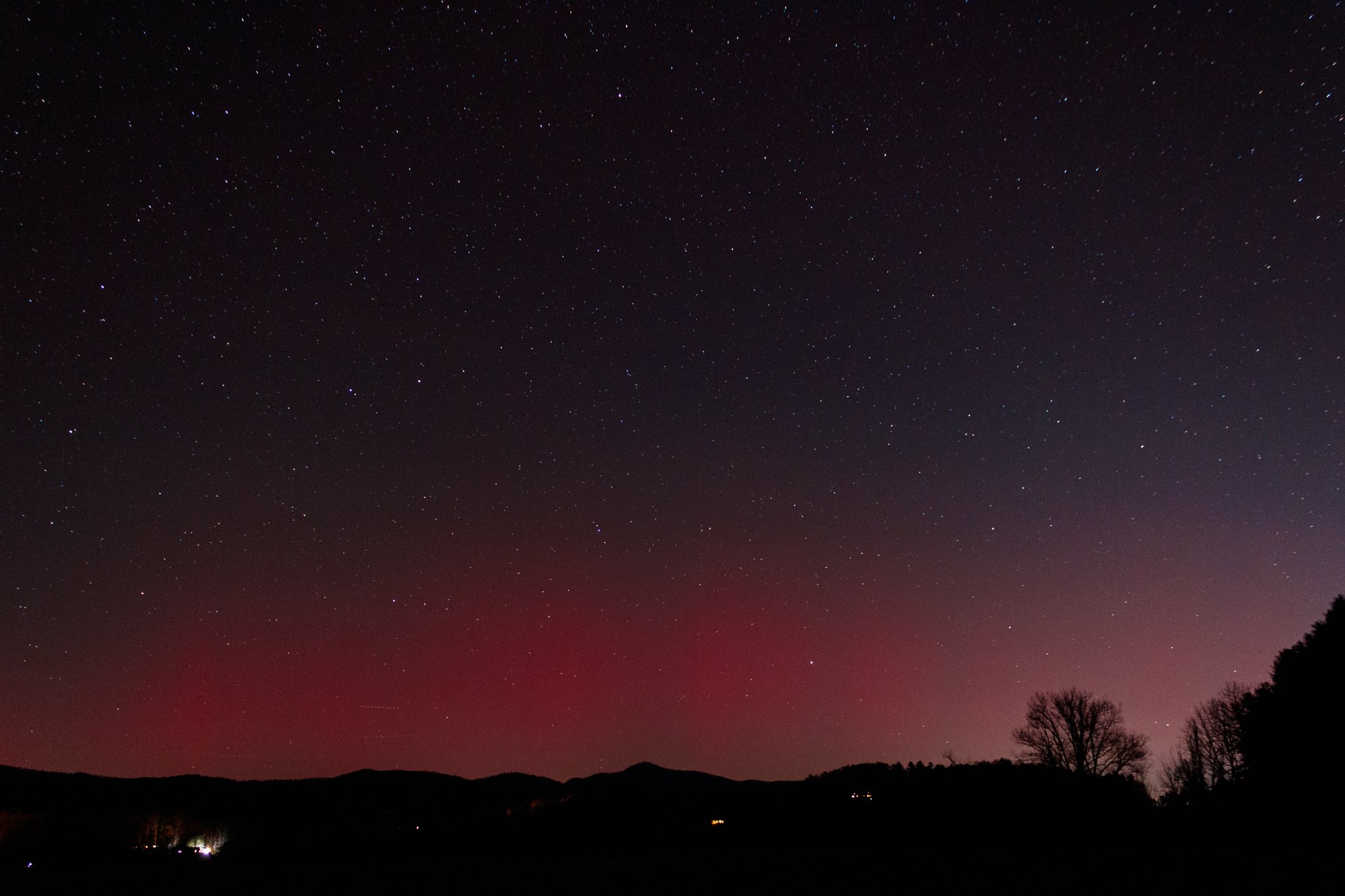 Nighttime landscape with a star-filled sky, distant hills, and silhouettes of trees.