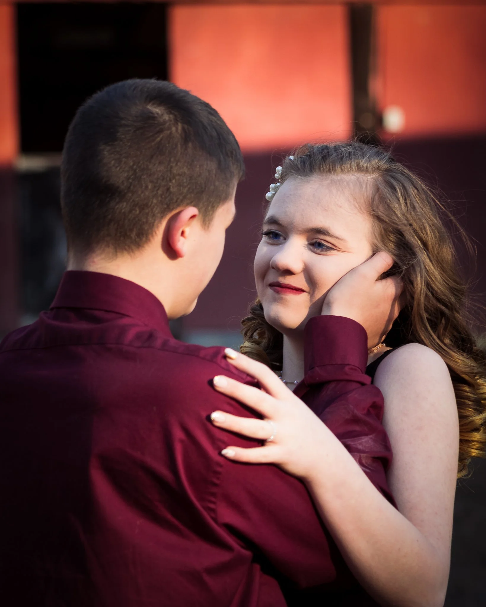 A young man and woman are dancing closely, gazing into each other's eyes, with the woman's hand resting on the man's shoulder and the man's hand gently holding her face.