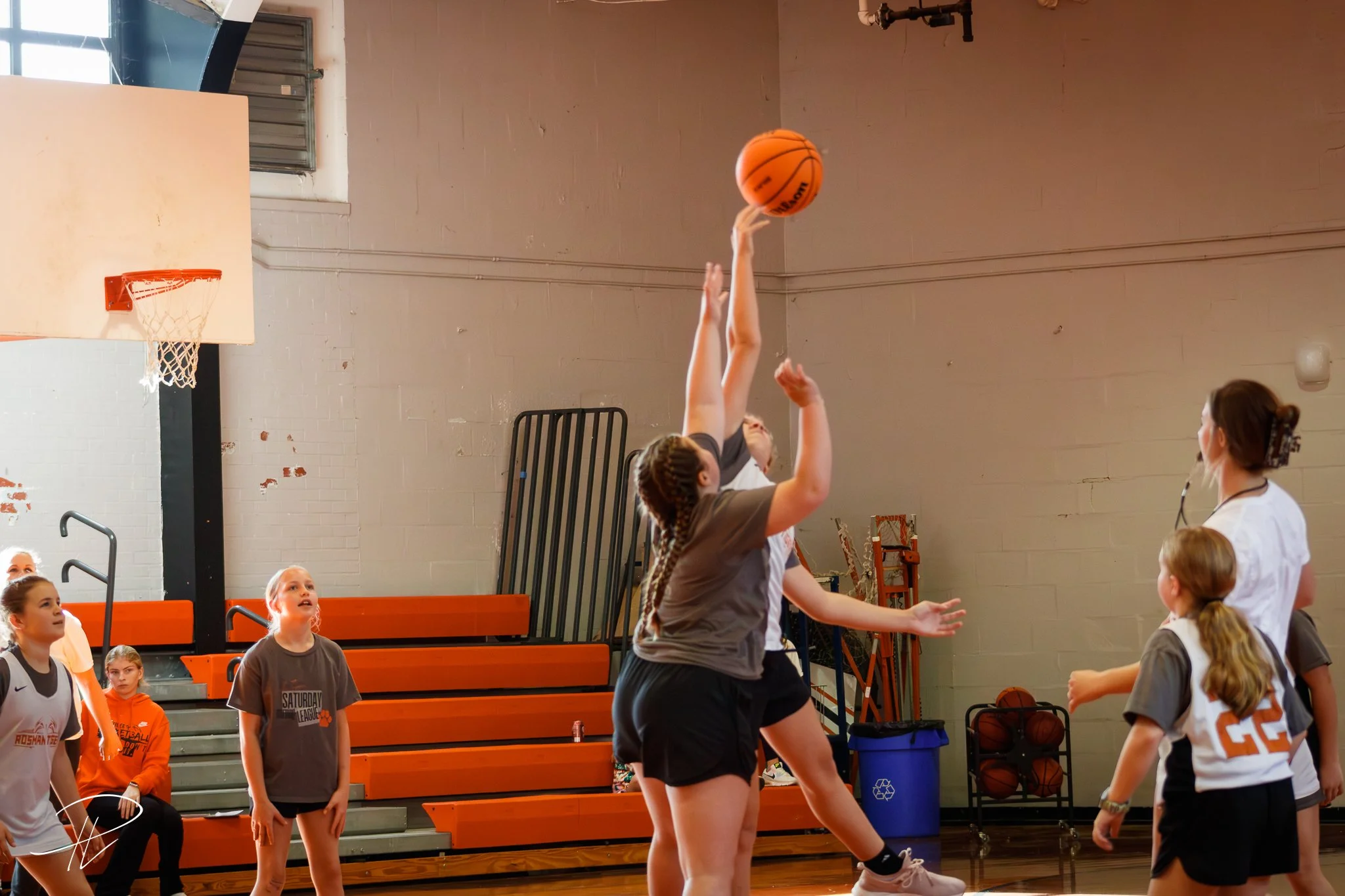 Young girls playing basketball in an indoor gym, with one girl shooting the ball towards the hoop as others watch.