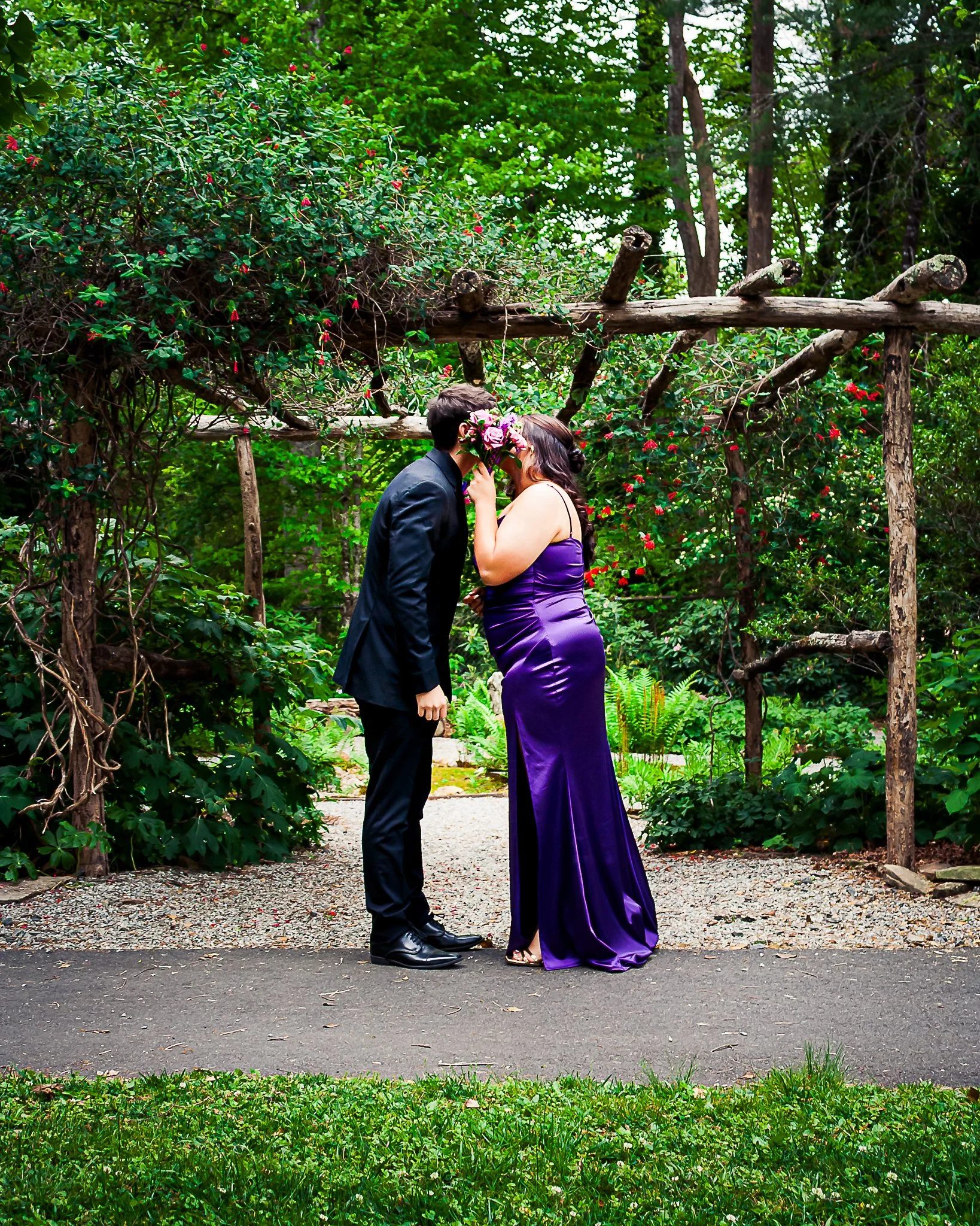 A couple dressed in formal attire sharing a kiss under a wooden archway in a lush garden setting, greenery surrounds them.