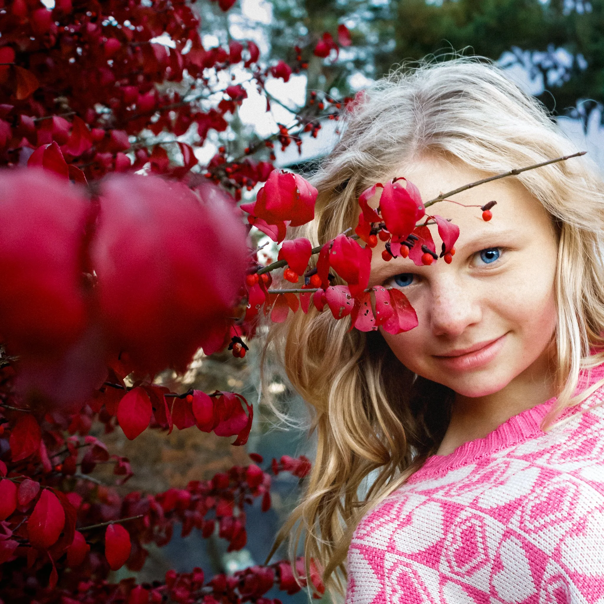 A young girl with blonde hair and blue eyes standing beside a red flowering bush, smiling softly.