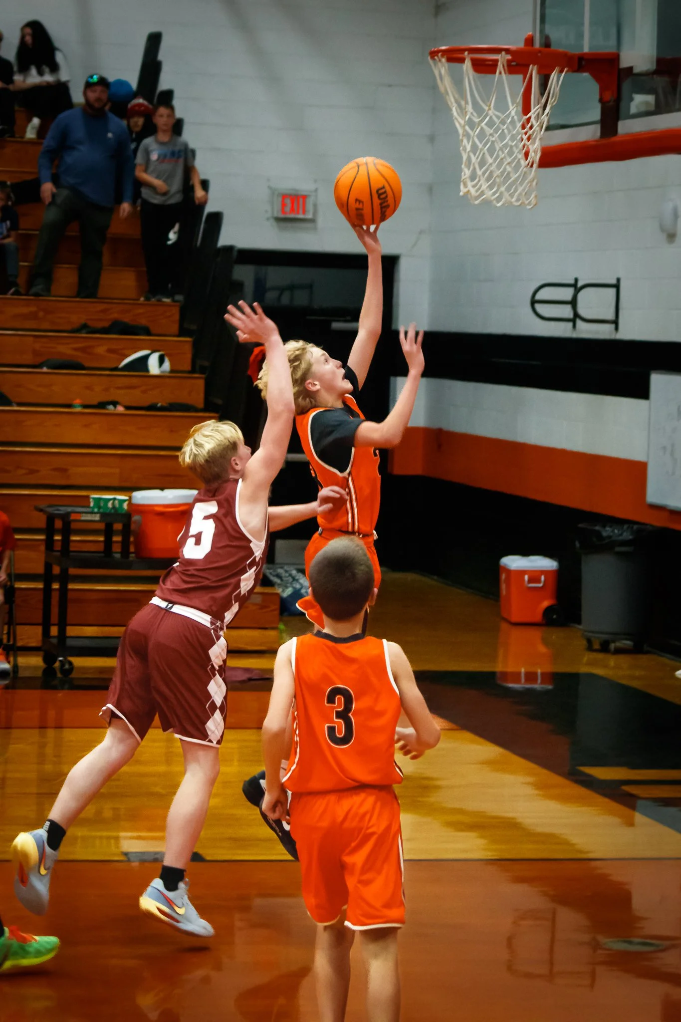 A young basketball player in an orange jersey is jumping, attempting to make a shot at the hoop. Two other kids are nearby, one in a maroon jersey and another in an orange jersey, watching the play. Spectators are visible in the background on bleache
