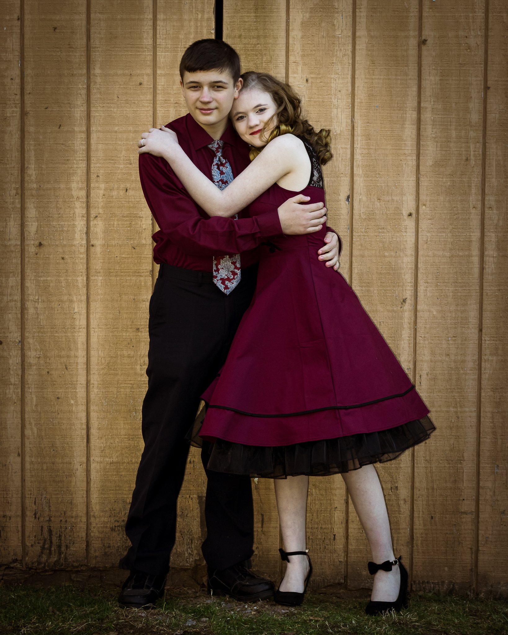 A young couple hugging in front of a wooden fence. The girl is wearing a maroon dress, and the boy is dressed in a maroon shirt with a patterned tie and black pants.