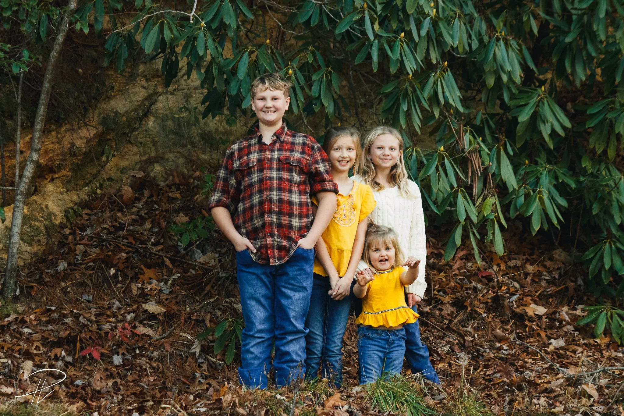 Four children standing outdoors among fallen leaves and green bushes, smiling at the camera.