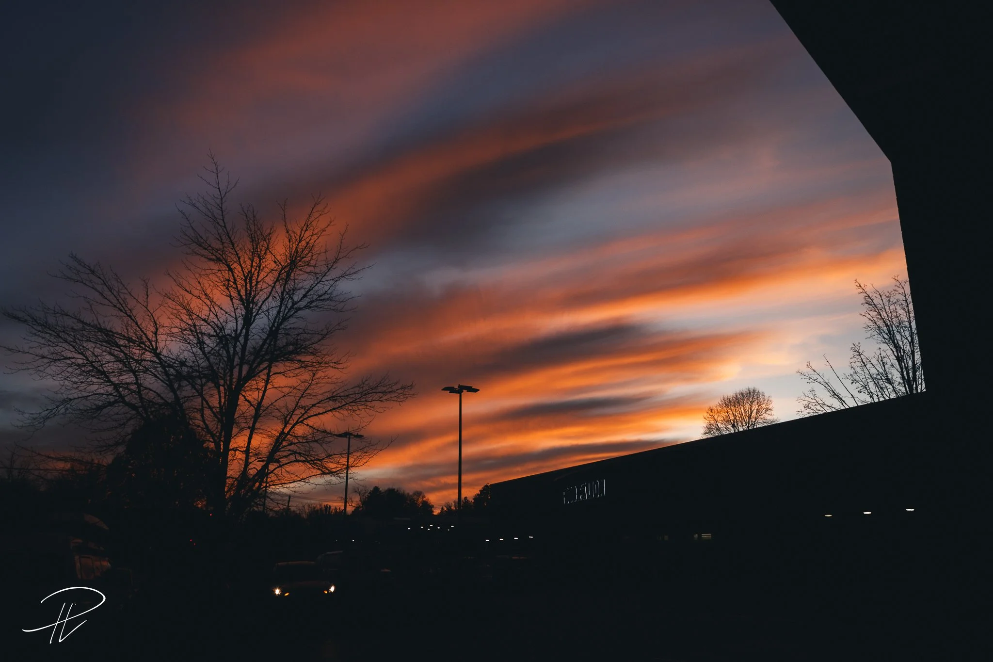 Sunset with vibrant orange and purple clouds in the sky, leafless trees silhouette, dark parking lot and building