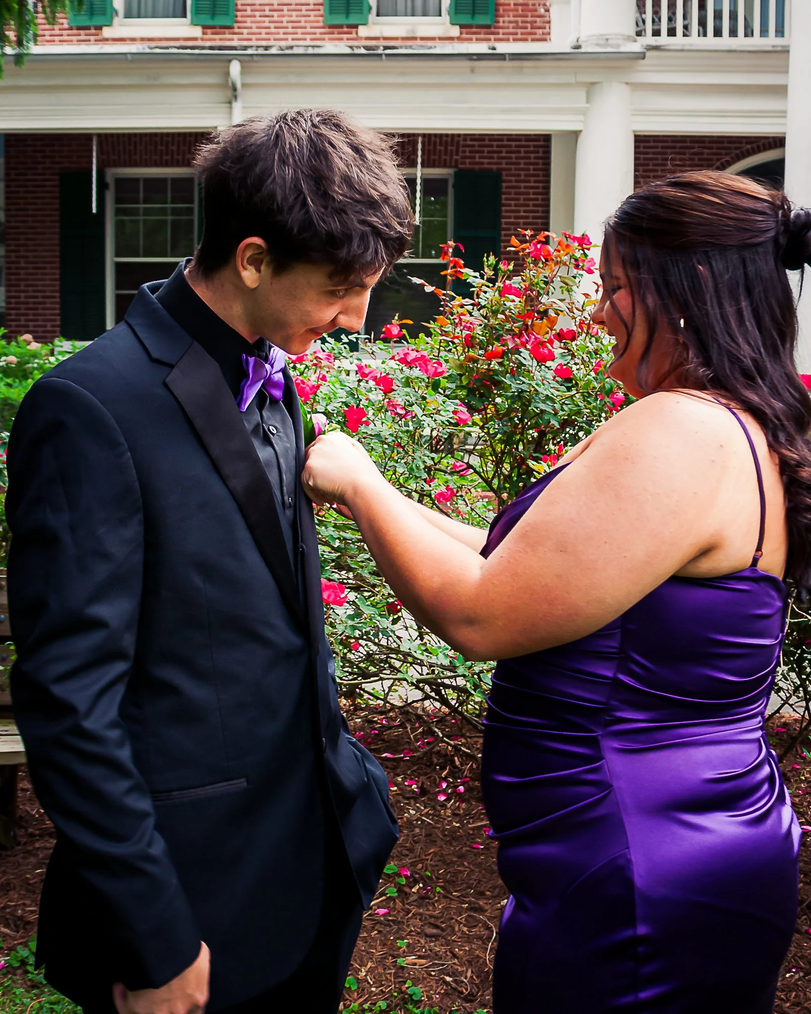 A couple dressed formally, the man in a tuxedo with a purple bow tie and the woman in a purple dress, are facing each other in a garden with pink flowers. The man is looking down at the woman's hand while she is touching his chest, both smiling.