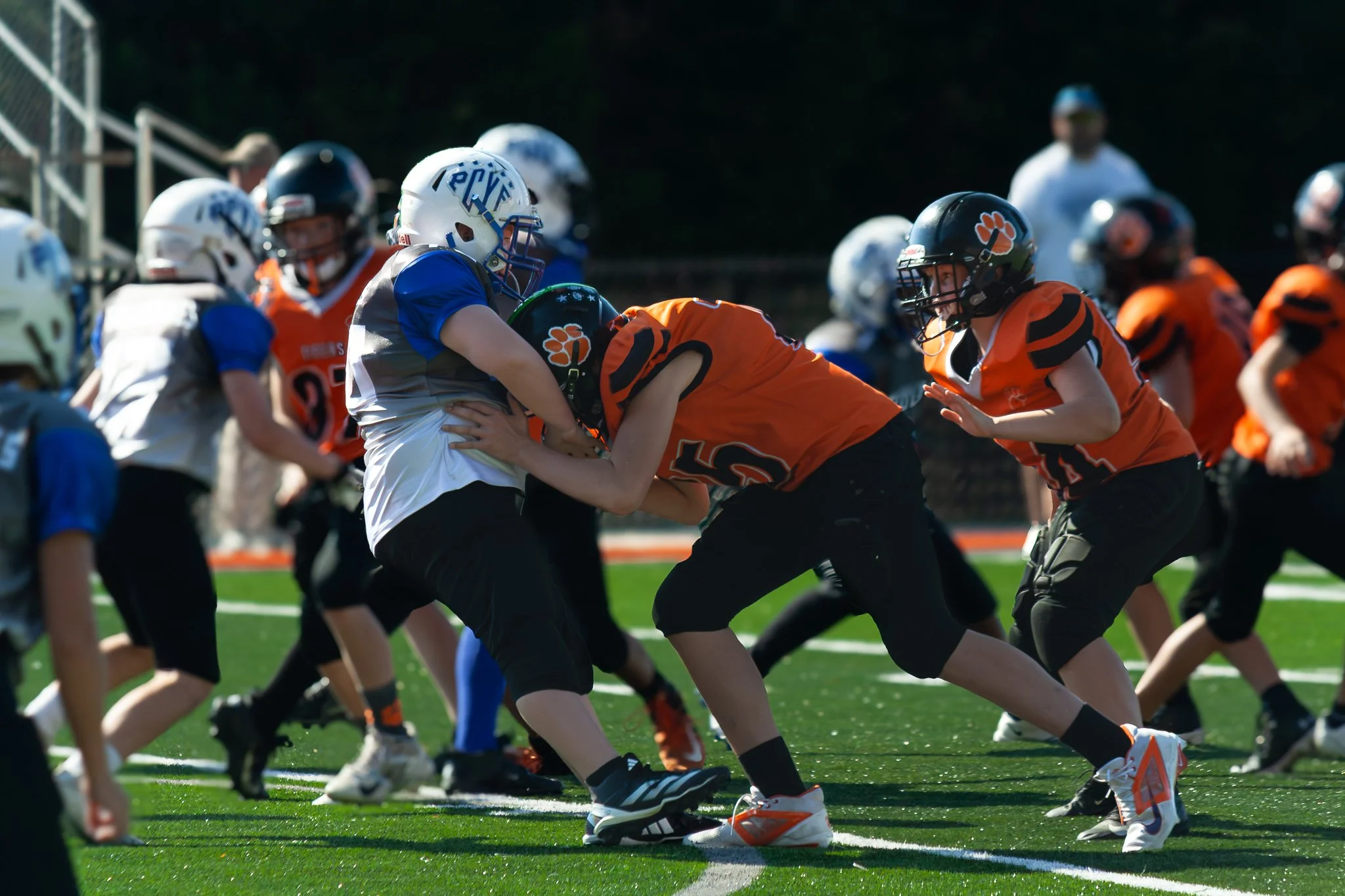 Youth football players in orange, white, and blue uniforms playing on a green field, with one player tackling another.