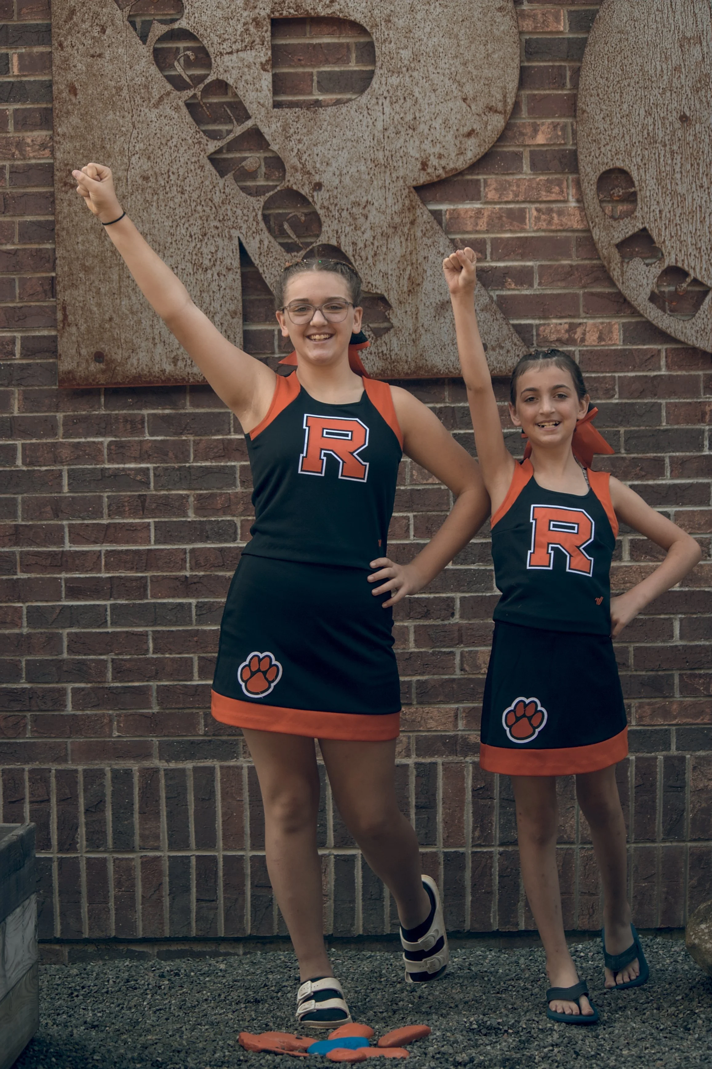 Two young girls in cheerleader uniforms with a large 'R' on the front, standing in front of a brick wall with a rusty metal sign, raising their arms in a cheer pose, smiling.
