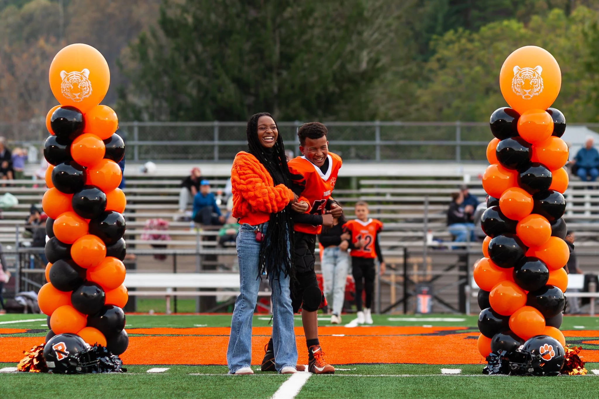 A woman and a young football player smiling and holding each other on a football field during a game event. There are orange and black balloons arranged on either side of the field entrance, and a football helmet with a tiger paw logo is on the groun