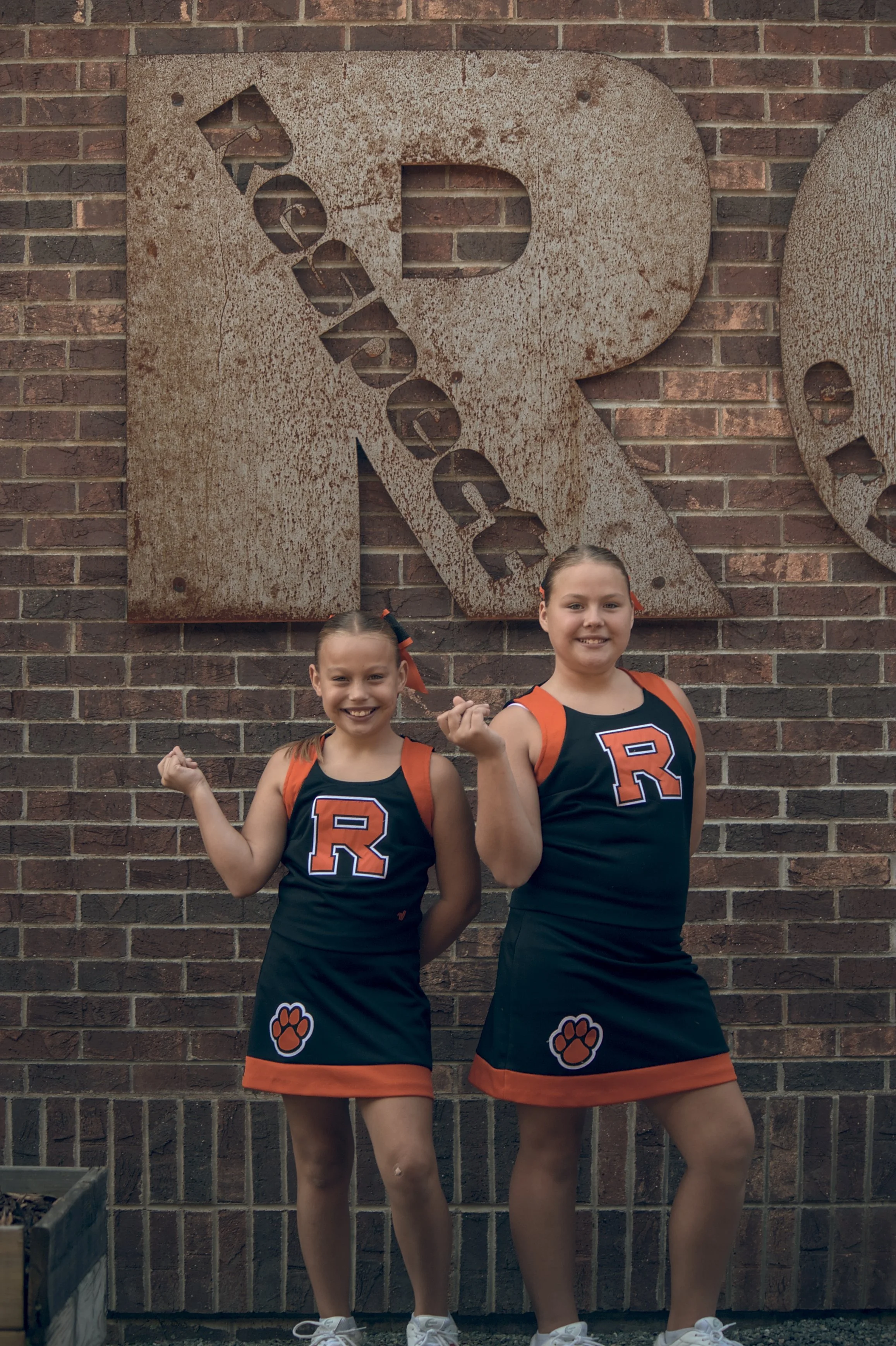 Two young cheerleaders in black and orange uniforms with a large letter R and a paw print on the front, standing in front of a brick wall with a large rusted letter R.