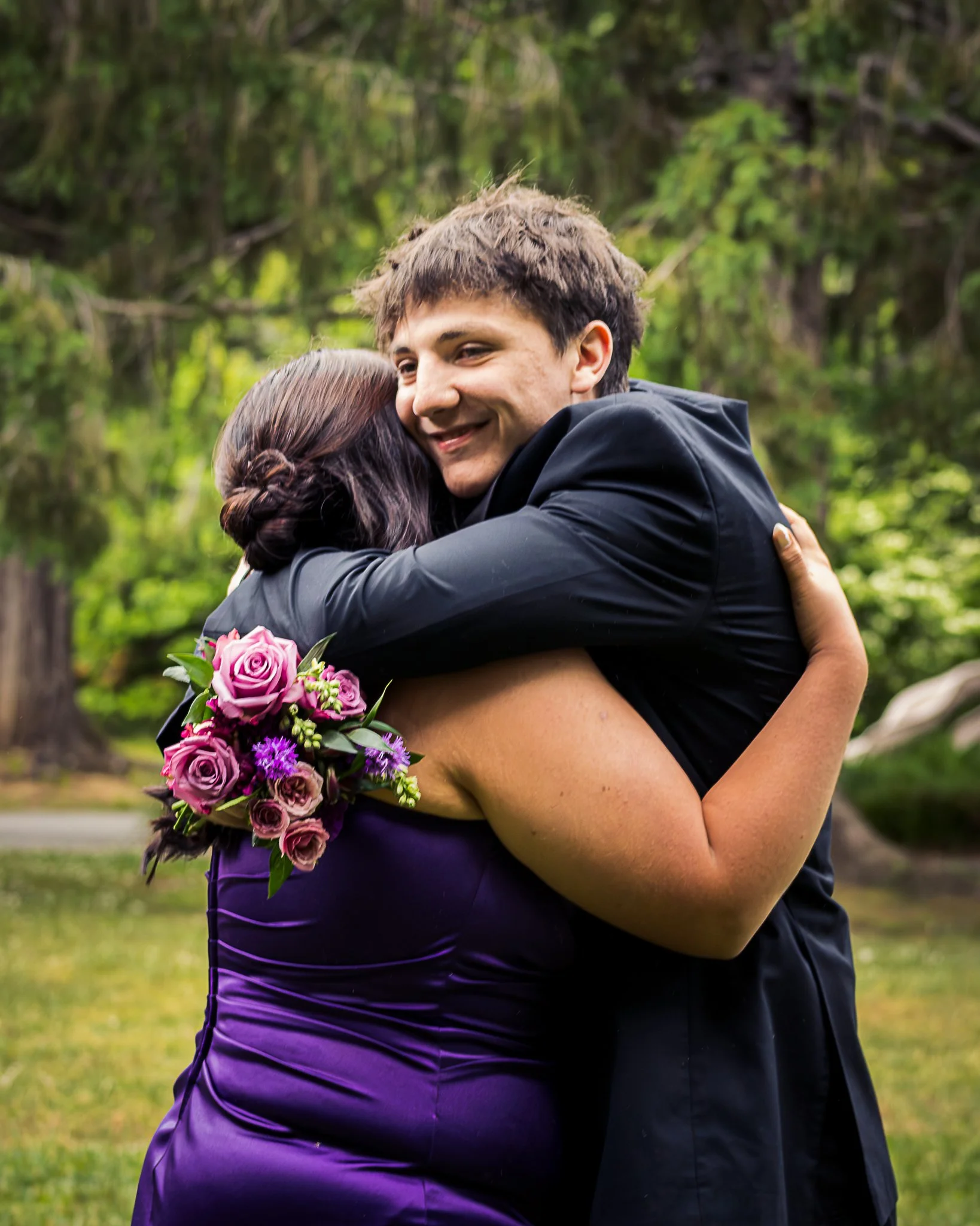 A man in a black suit hugging a woman in a purple dress, holding a bouquet of purple and pink roses, outdoors in a park with greenery in the background.
