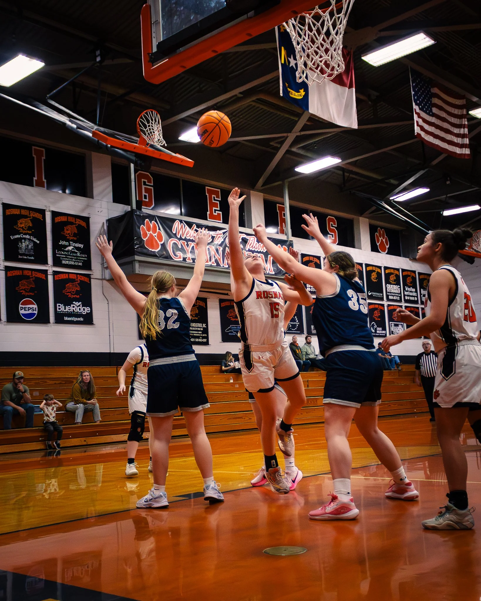 Girls playing basketball in indoor gymnasium, with some players jumping and reaching for the ball near the hoop. Spectators sitting on wooden bleachers in the background.