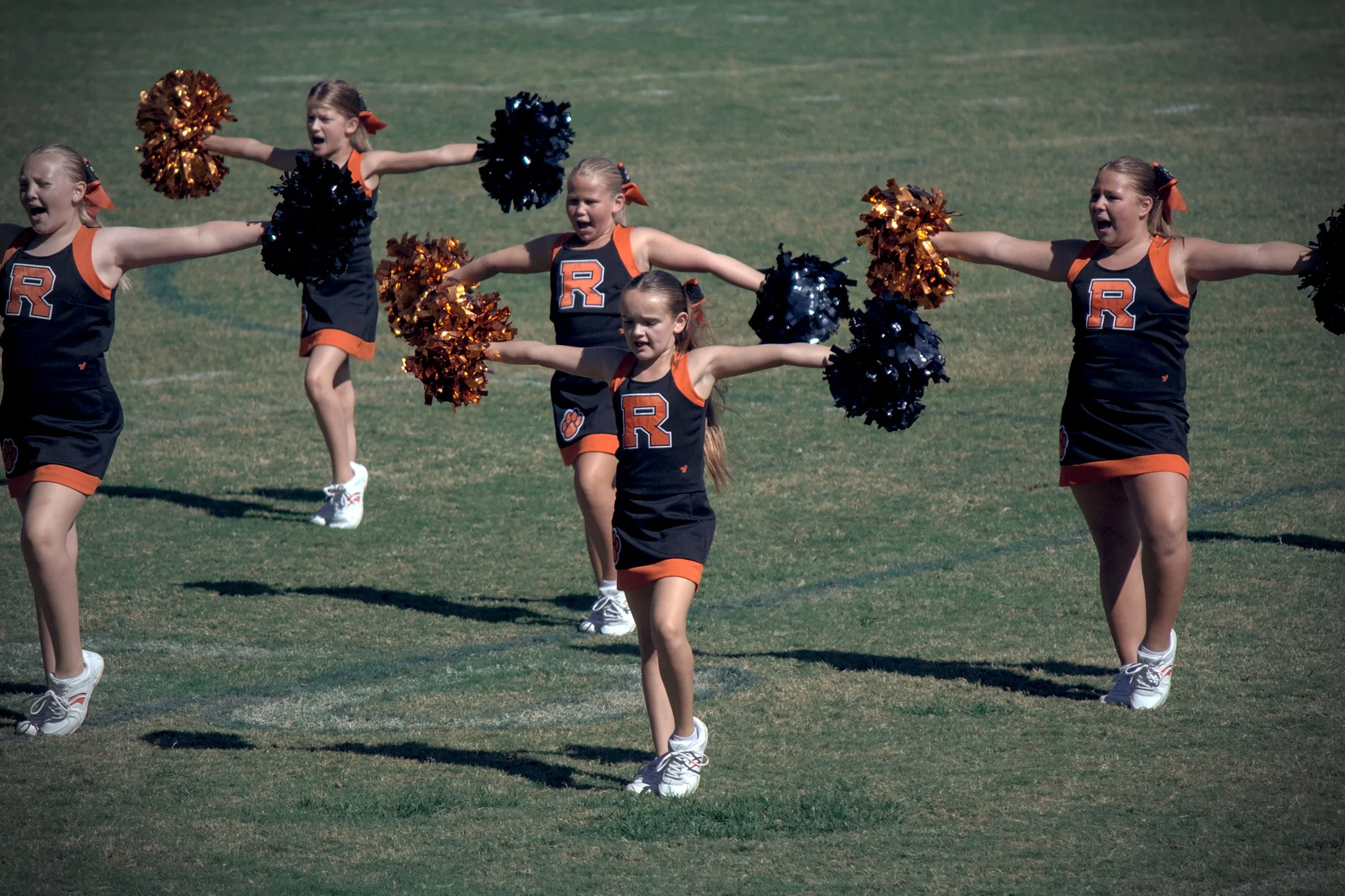 Group of young cheerleaders performing on a grassy field, wearing black and orange uniforms with a large letter 'R' on the front, holding pom-poms in black and orange, with their arms extended outward.