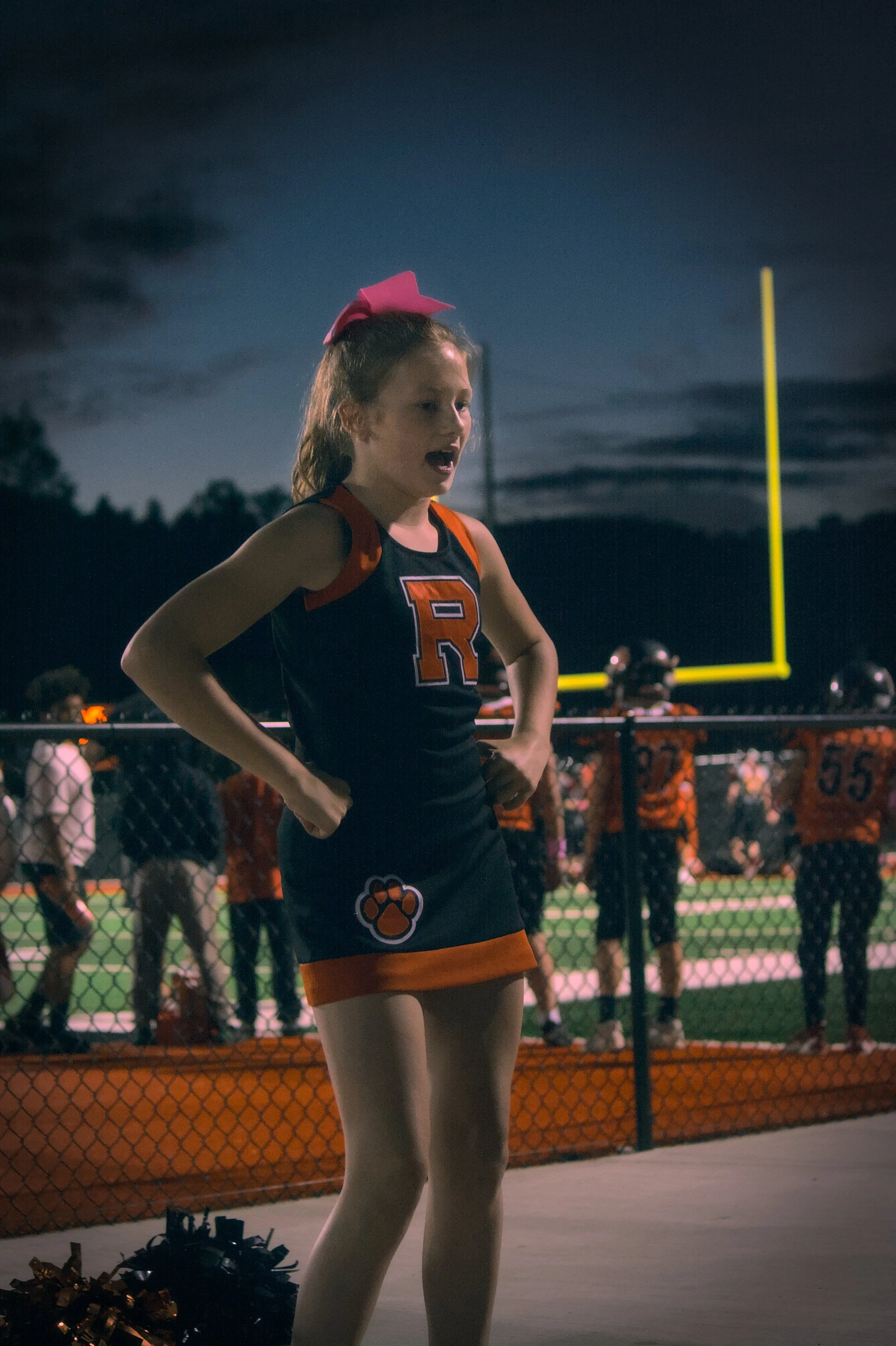 A young female cheerleader wearing a black and orange cheerleading uniform with a pink bow on her hair, standing with her hands on her hips on a football field during night time.