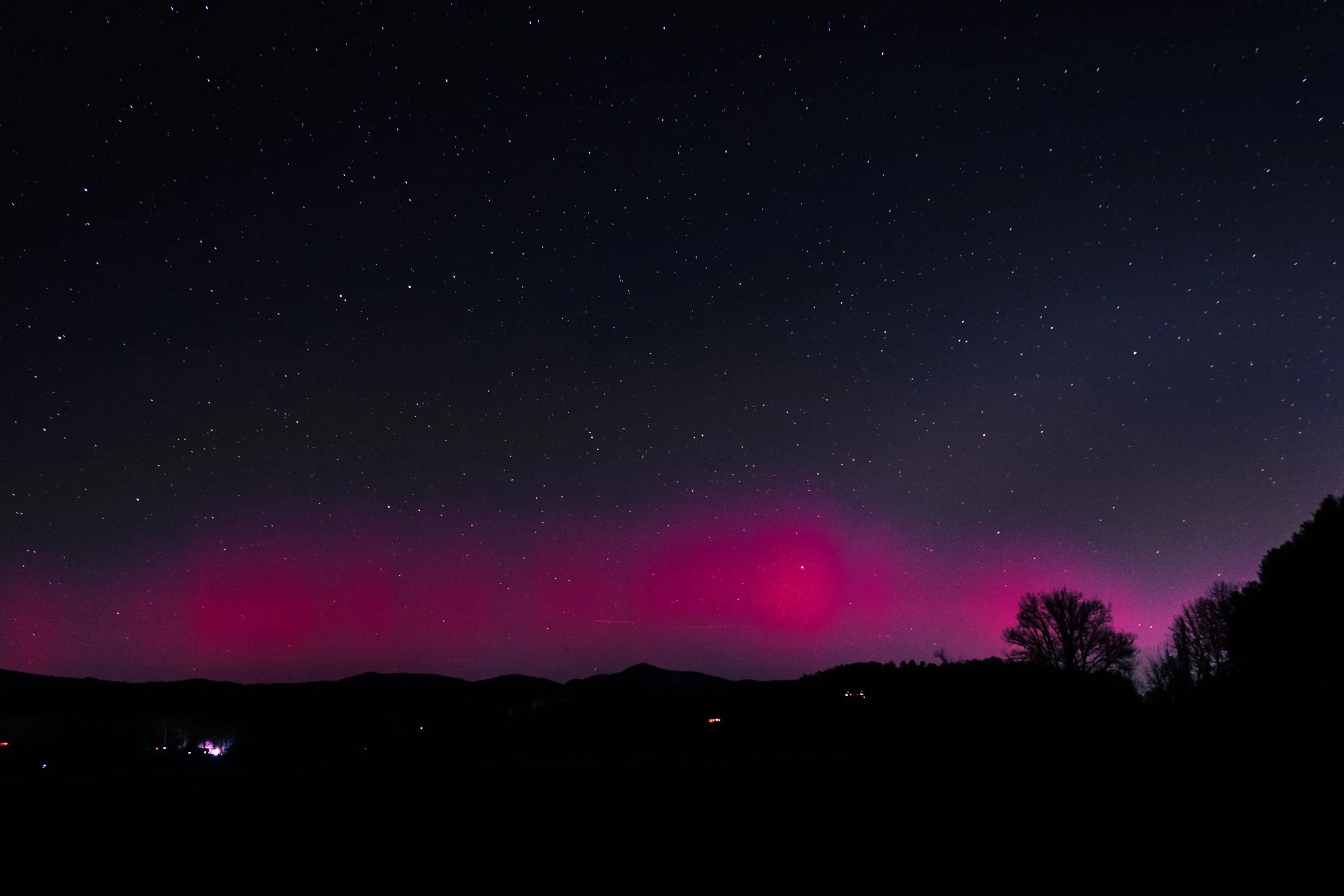 Night sky with stars and pink aurora over a silhouette of trees and mountains.