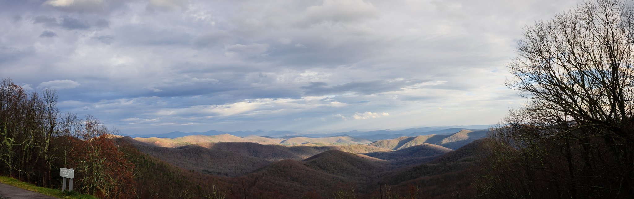 Panoramic view of rolling mountains with a cloudy sky overhead, some trees in the foreground, and a road sign on the bottom left.
