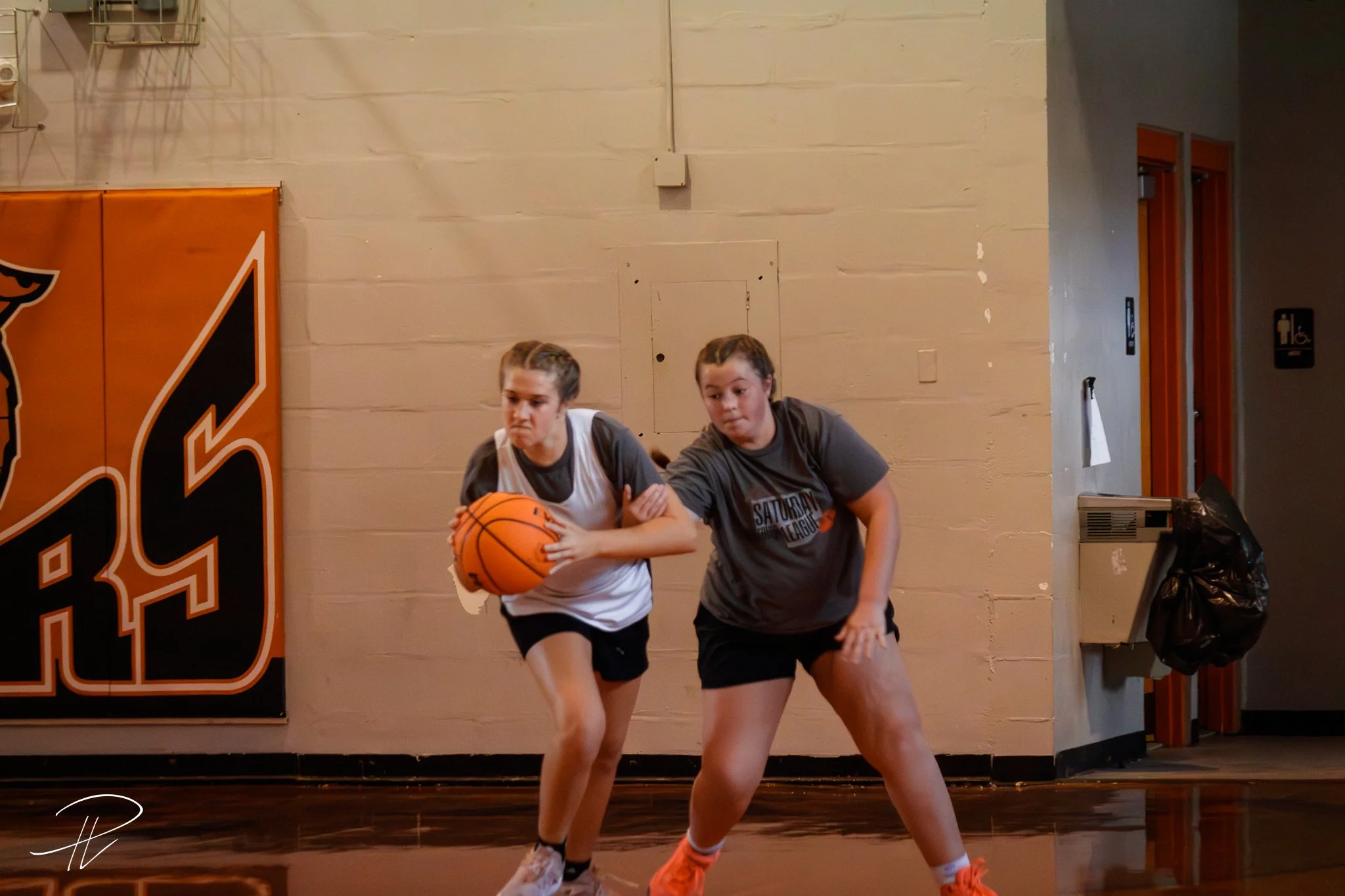 Two young female basketball players practicing on an indoor court, with one player dribbling the ball while the other defends, against a beige wall with a large orange and black banner.