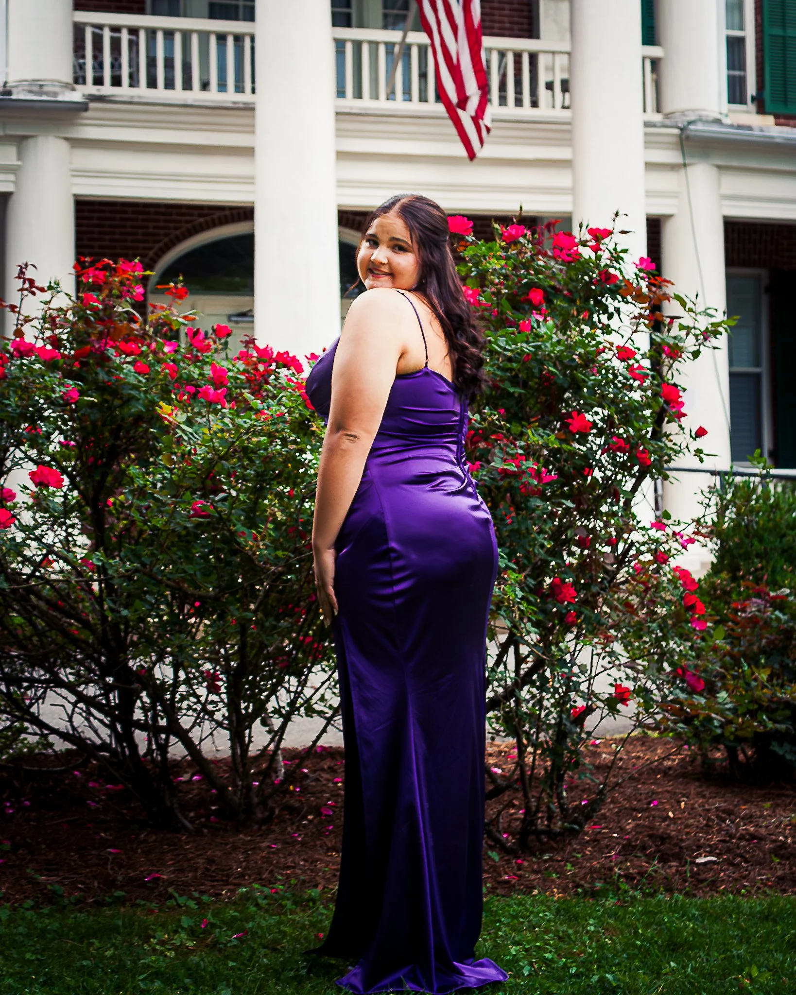 A woman in a purple satin dress standing outdoors in front of pink flowers and a house with white columns, green shutters, and an American flag hanging from the balcony.