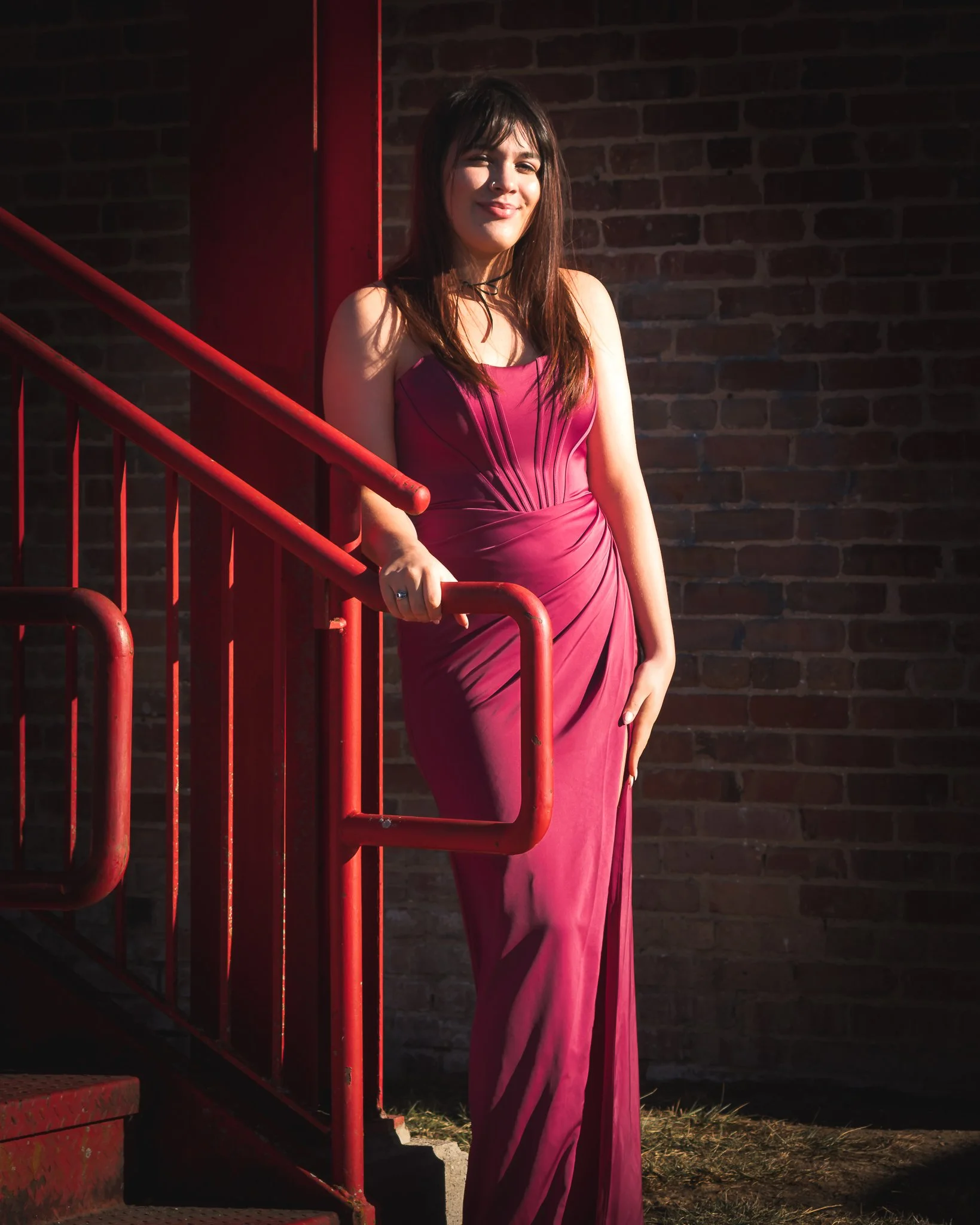 A young woman in a long pink dress standing next to a red metal railing outside, with a brick wall in the background.