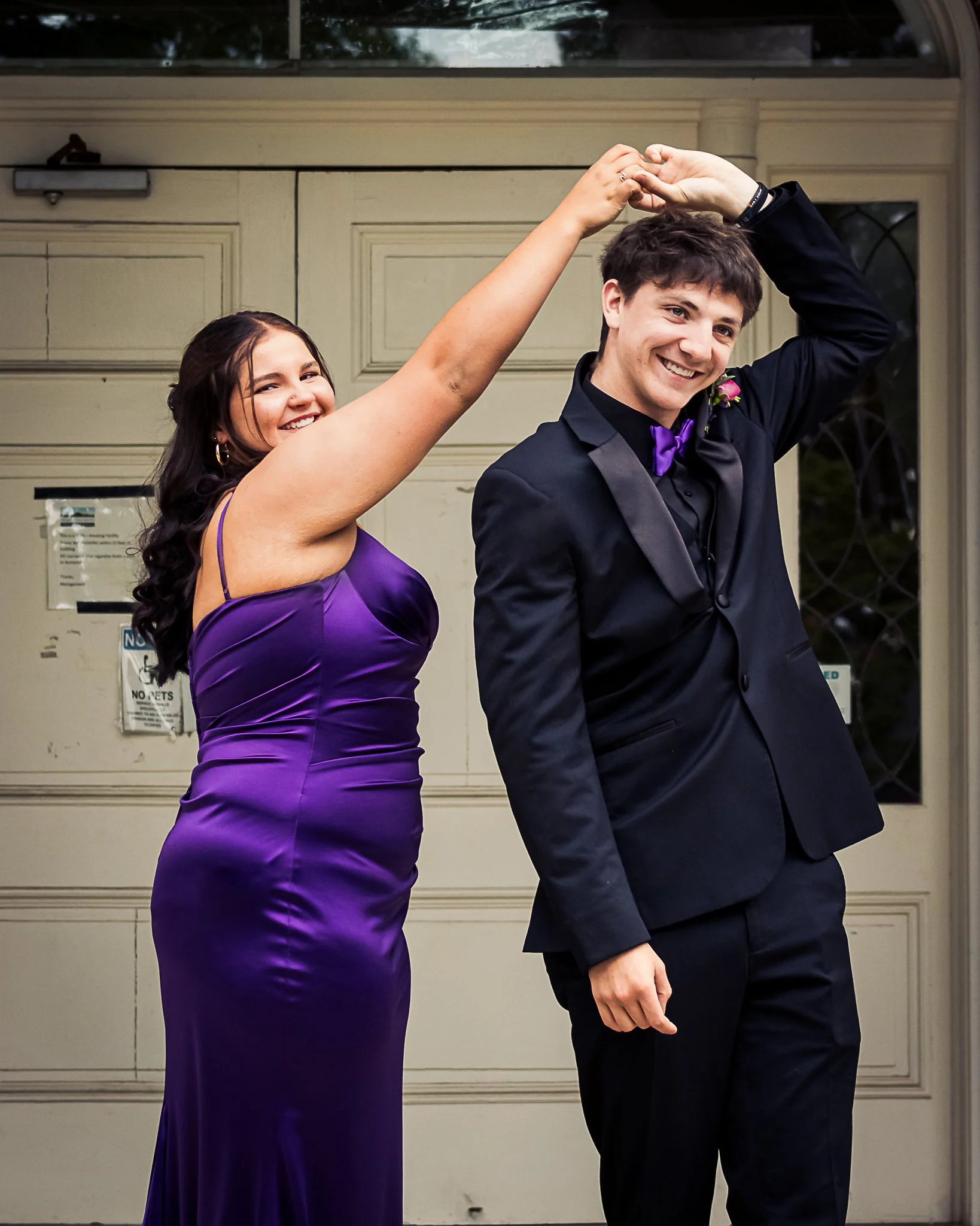 Two young adults, one woman and one man, dressed in formal attire, dancing together outside in front of a beige garage door. The woman wears a purple dress, and the man wears a black tuxedo with a purple bow tie, smiling while holding hands and twirl