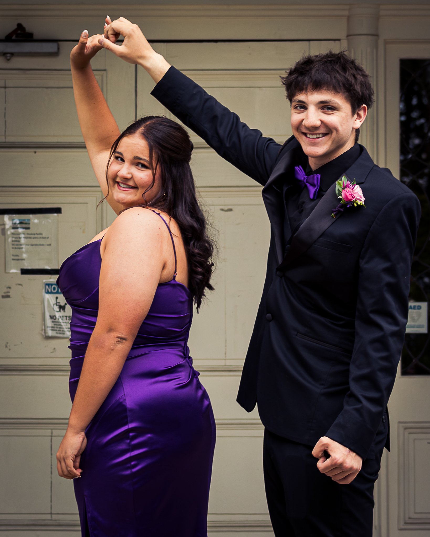 A woman in a purple dress and a man in a black tuxedo dancing together, holding hands, and smiling outdoors in front of a building.