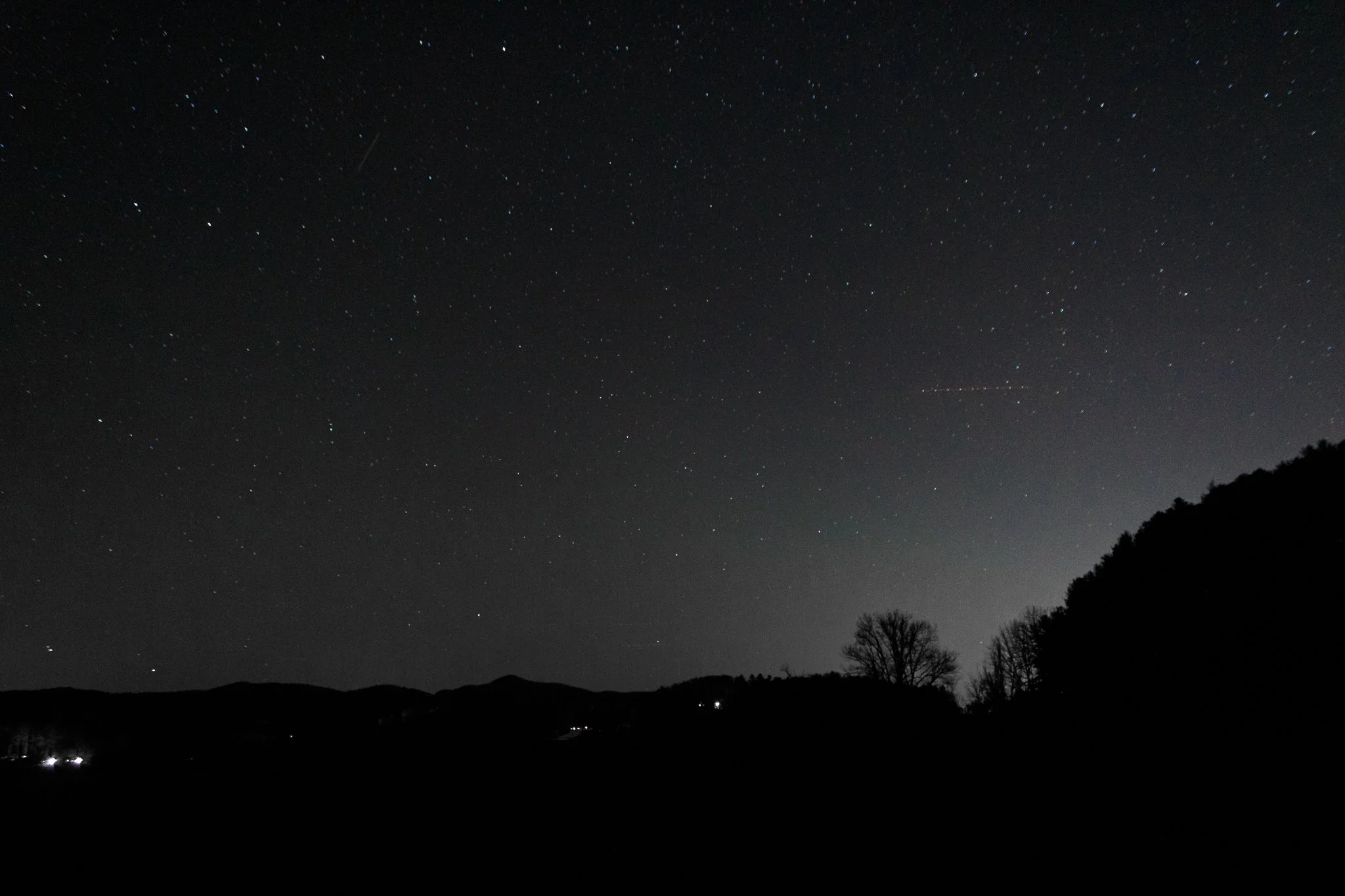 Night sky filled with stars over a silhouetted hilly landscape and trees.