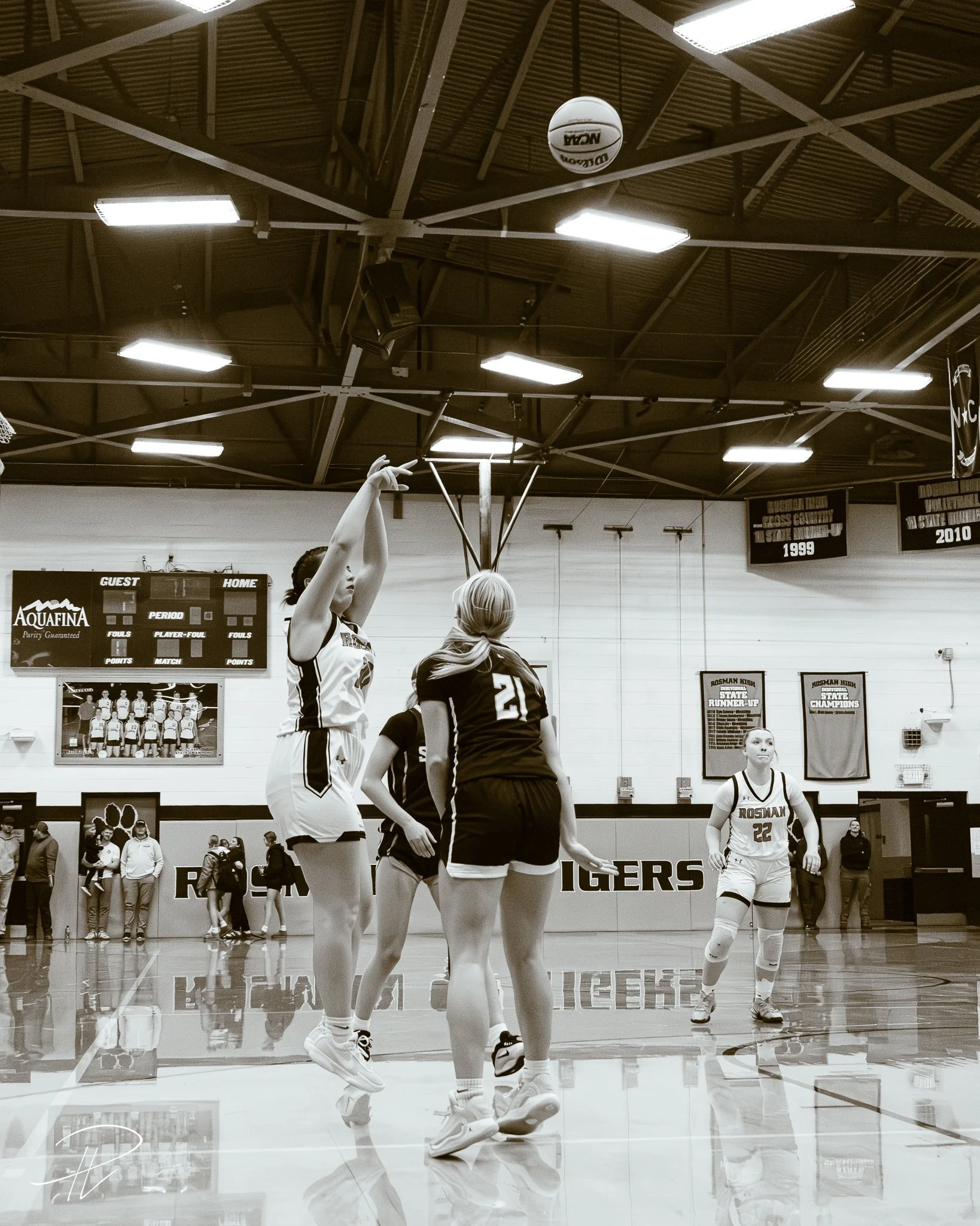 A girls' basketball game in a gymnasium with players jumping to catch or block a basketball. Two players are in focus, one jumping with arms extended, the other with her back to the camera. The gym has banners on the wall and a scoreboard in the back