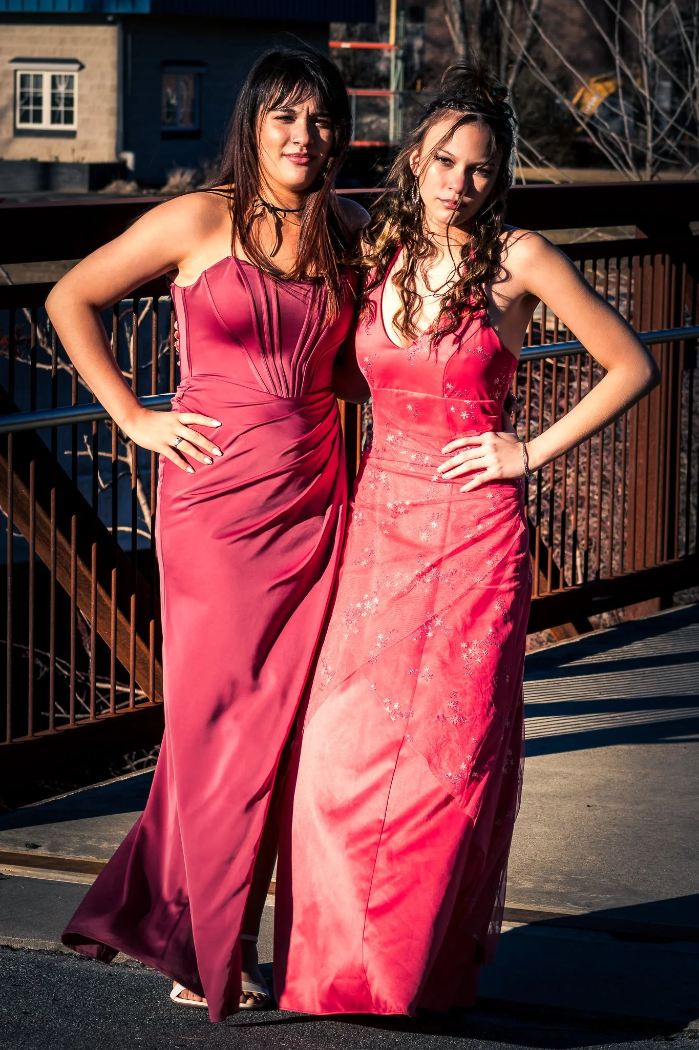 Two women in pink evening gowns standing outdoors on a bridge with a cityscape background.