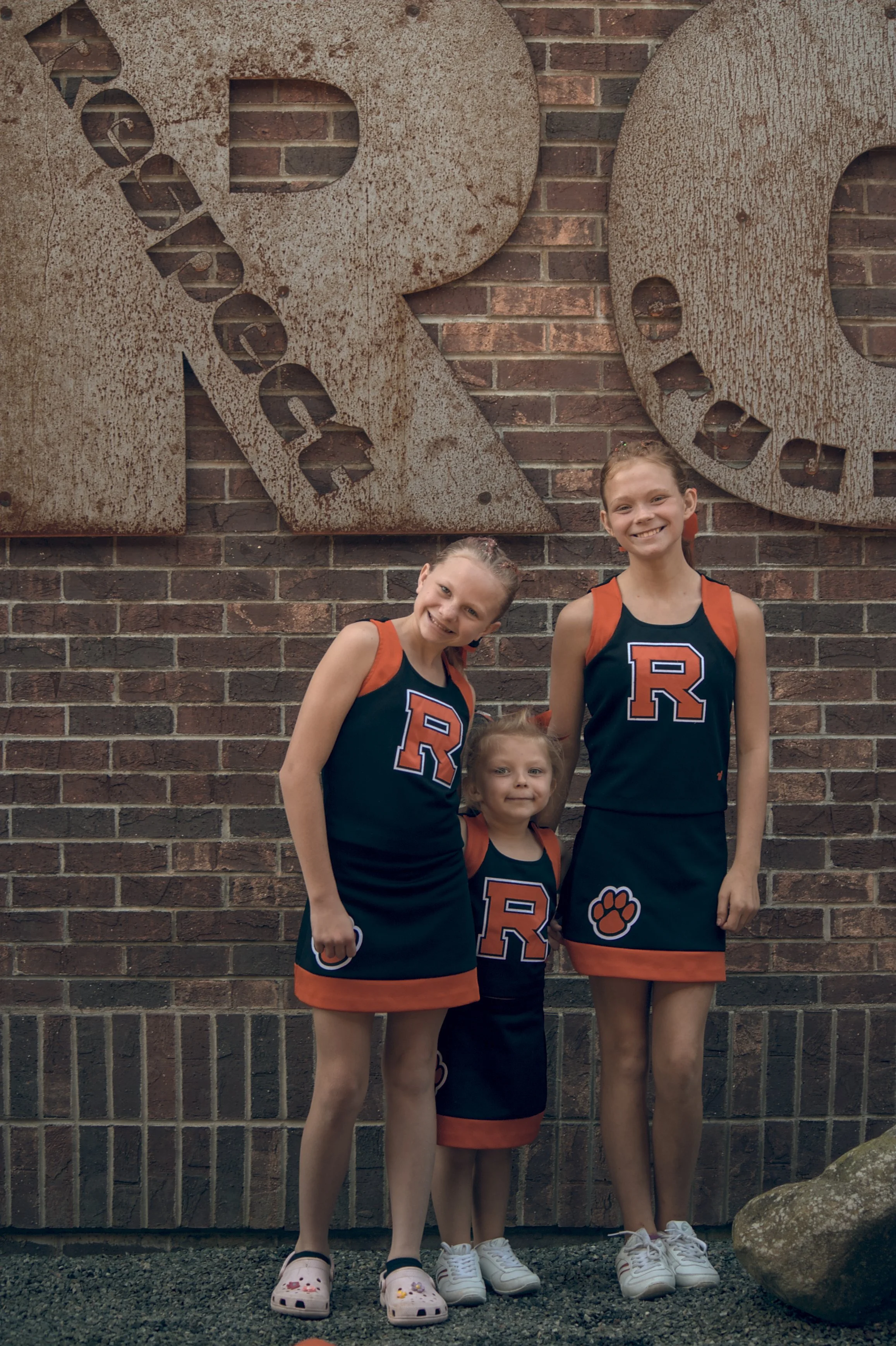 Three young girls in matching cheerleading uniforms standing in front of a brick wall with large, rusty metal letters and words on it. The girls are smiling.