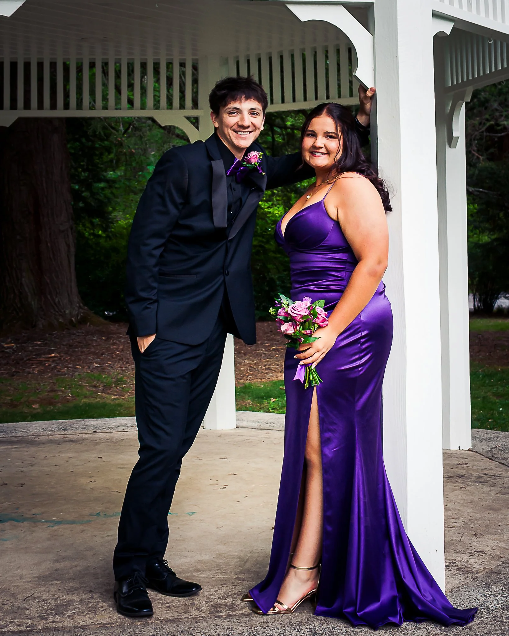 A smiling young man in a black tuxedo with a purple bowtie and pink boutonniere standing next to a smiling young woman in a purple satin gown with a thigh-high slit, holding a bouquet of pink roses. They are posing under a white gazebo with a wooded 