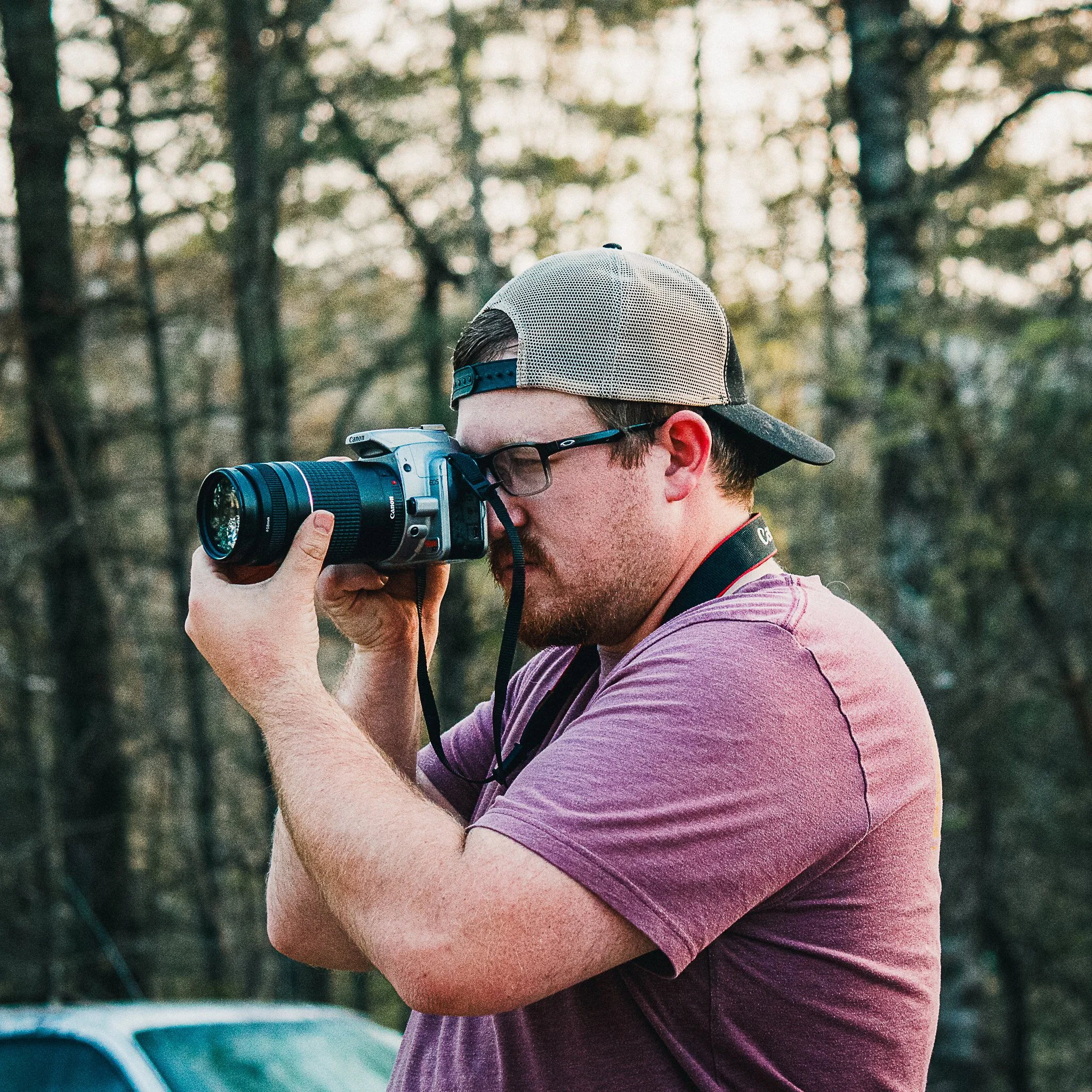 Person with a beard and glasses taking a photograph outdoors in a wooded area, wearing a baseball cap and a purple shirt.