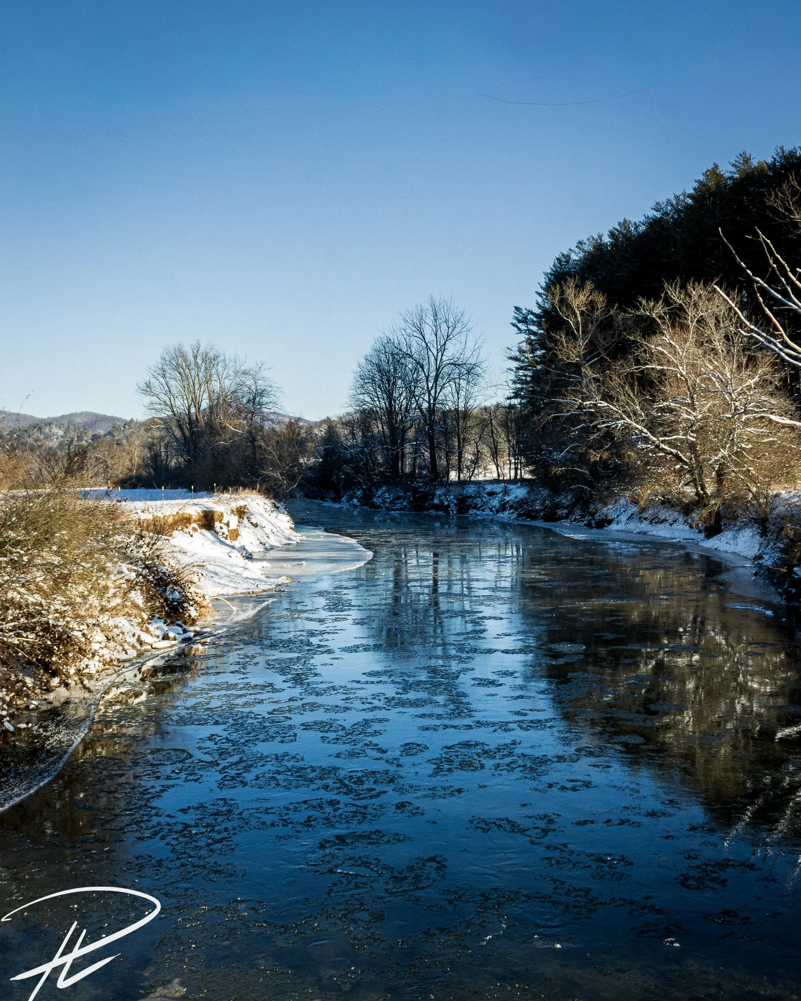 A winter landscape featuring a partially frozen river with ice patches and snow-covered banks, surrounded by leafless trees under a clear blue sky.