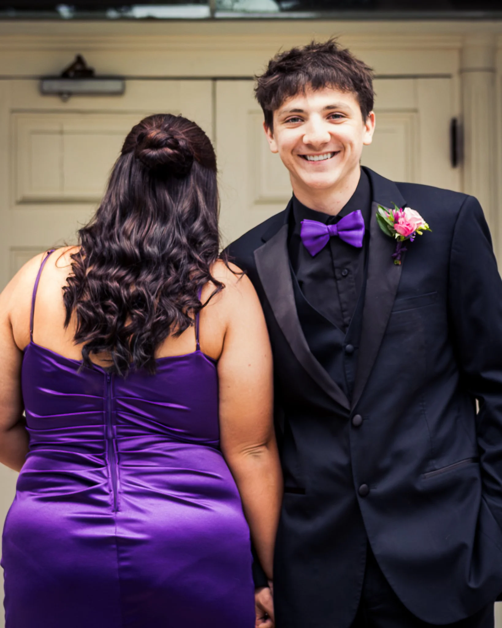 A smiling young man in a tuxedo with a purple bow tie and boutonnière, standing next to a woman in a purple dress, at a formal event with beige-background paneling.