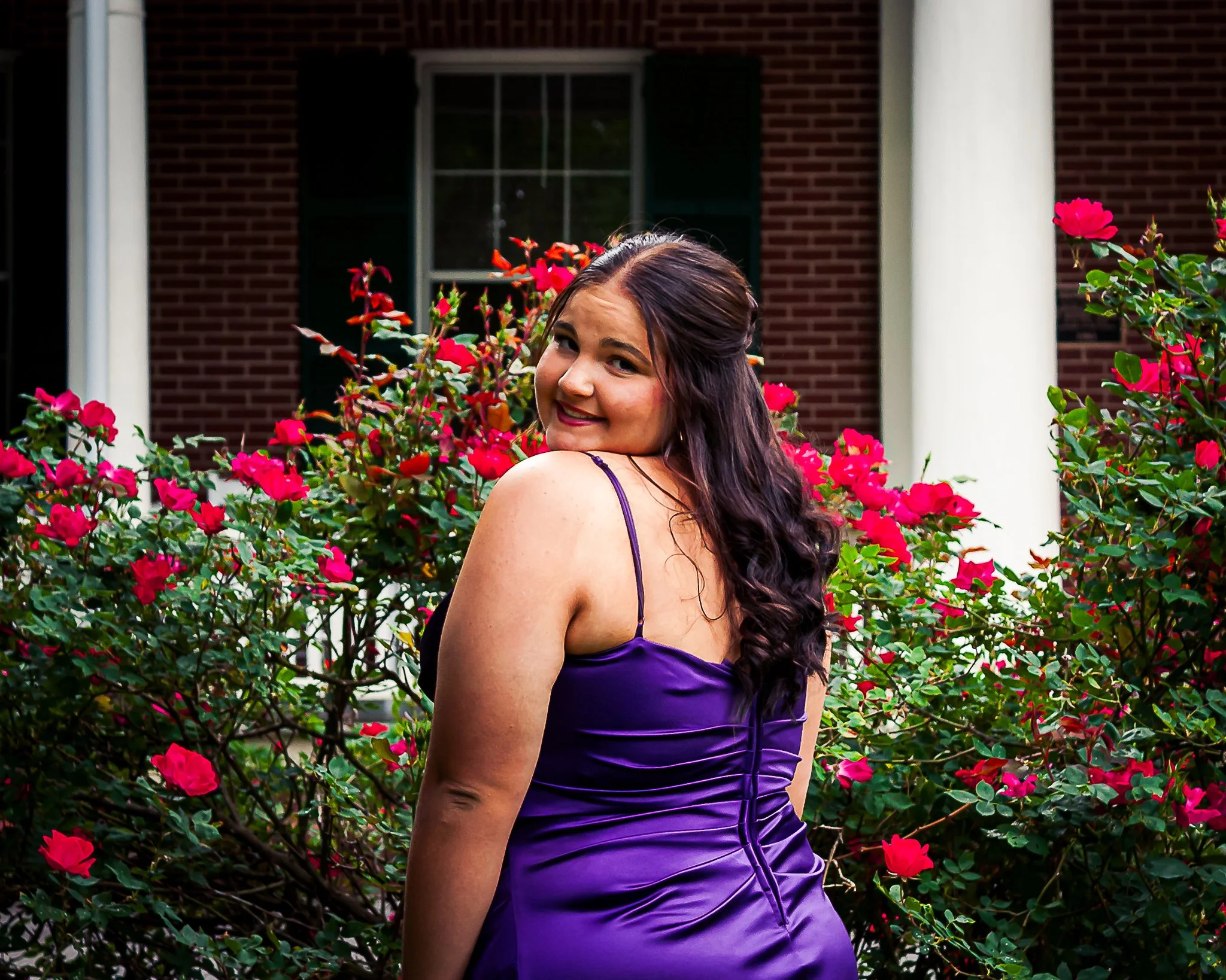 A woman with long dark hair and a purple dress is standing among bright pink flowers in front of a brick house with white columns and a window.