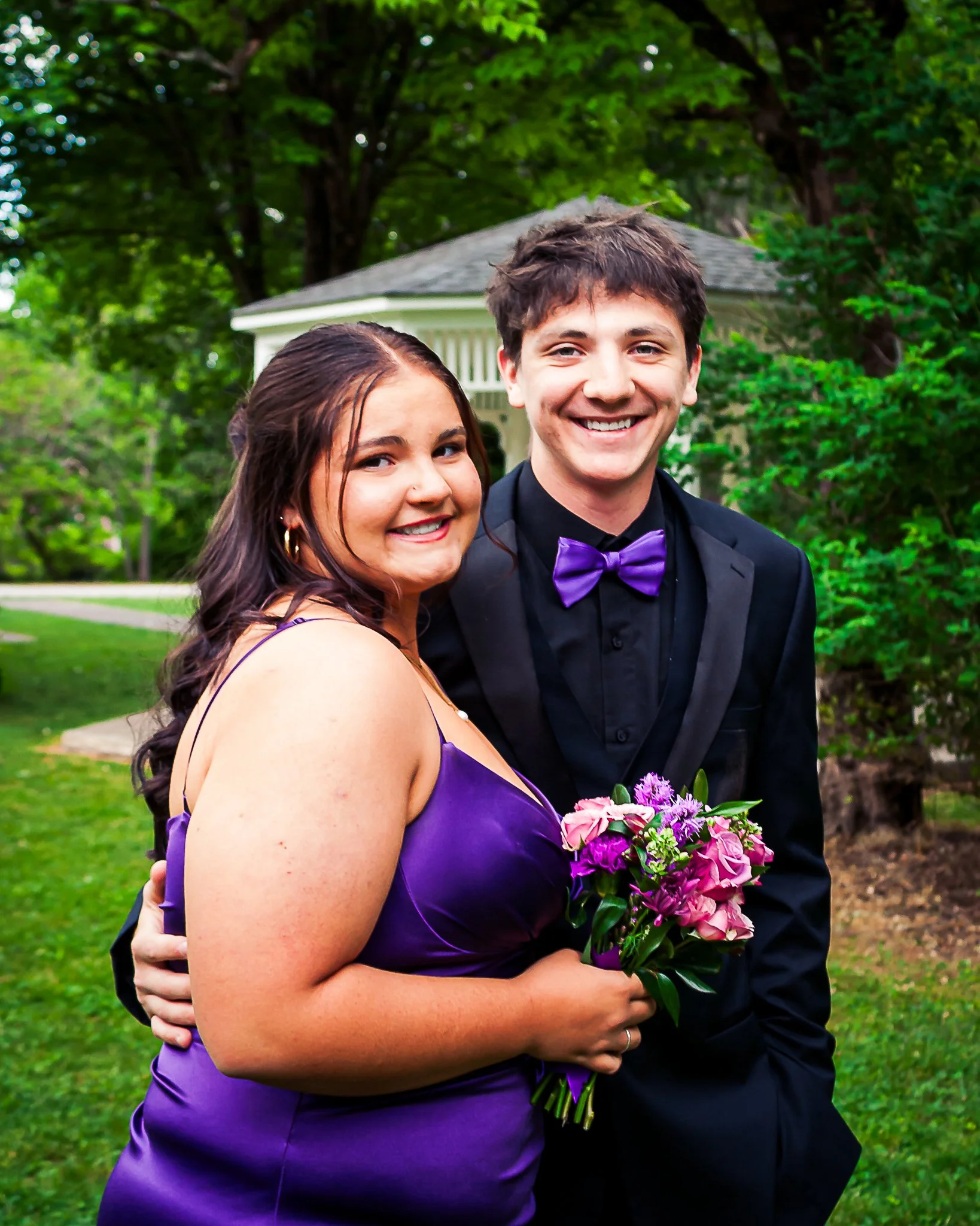 A smiling couple embracing outdoors, the woman in a purple dress holding a bouquet of pink and purple flowers, and the man in a black tuxedo with a purple bow tie, with greenery and trees in the background.